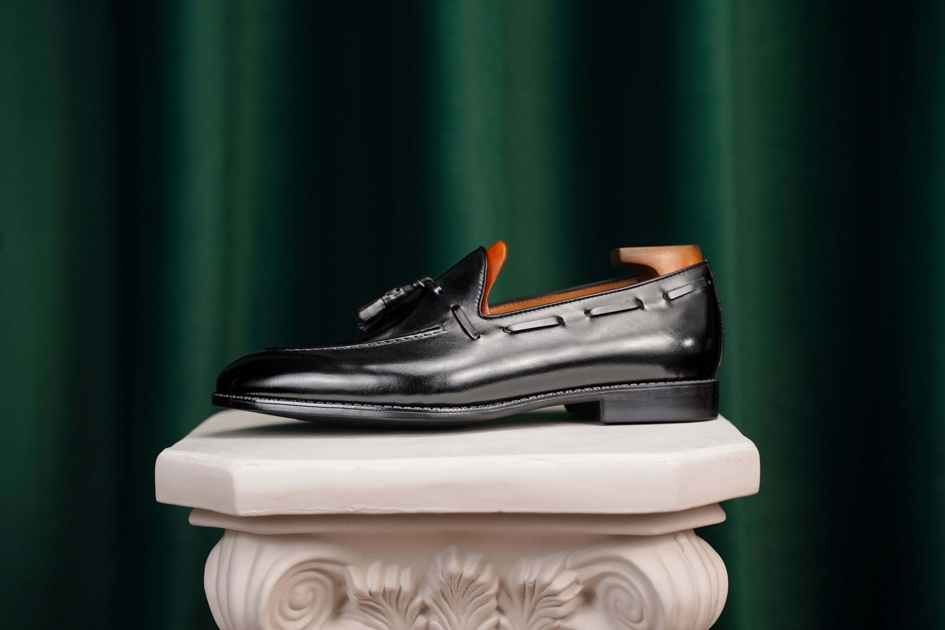 HolloShoe Handmade Black Tassel Leather Loafers – side view with shoe tree, resting on a white classical pedestal, highlighting polished black leather for refined impact.