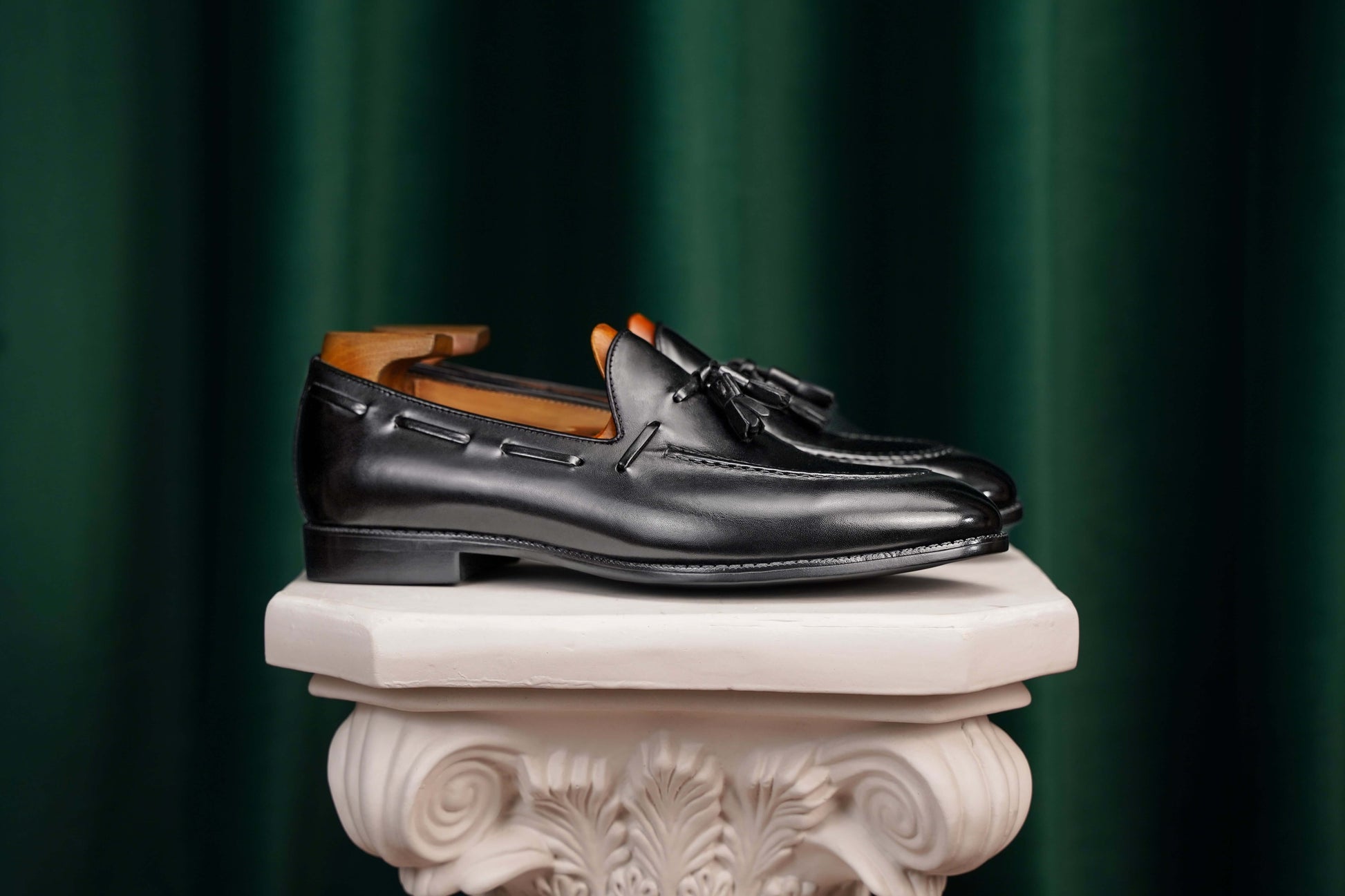 HolloShoe Handmade Black Tassel Leather Loafers – side view with shoe tree, resting on a white classical pedestal, highlighting polished black leather for refined impact.