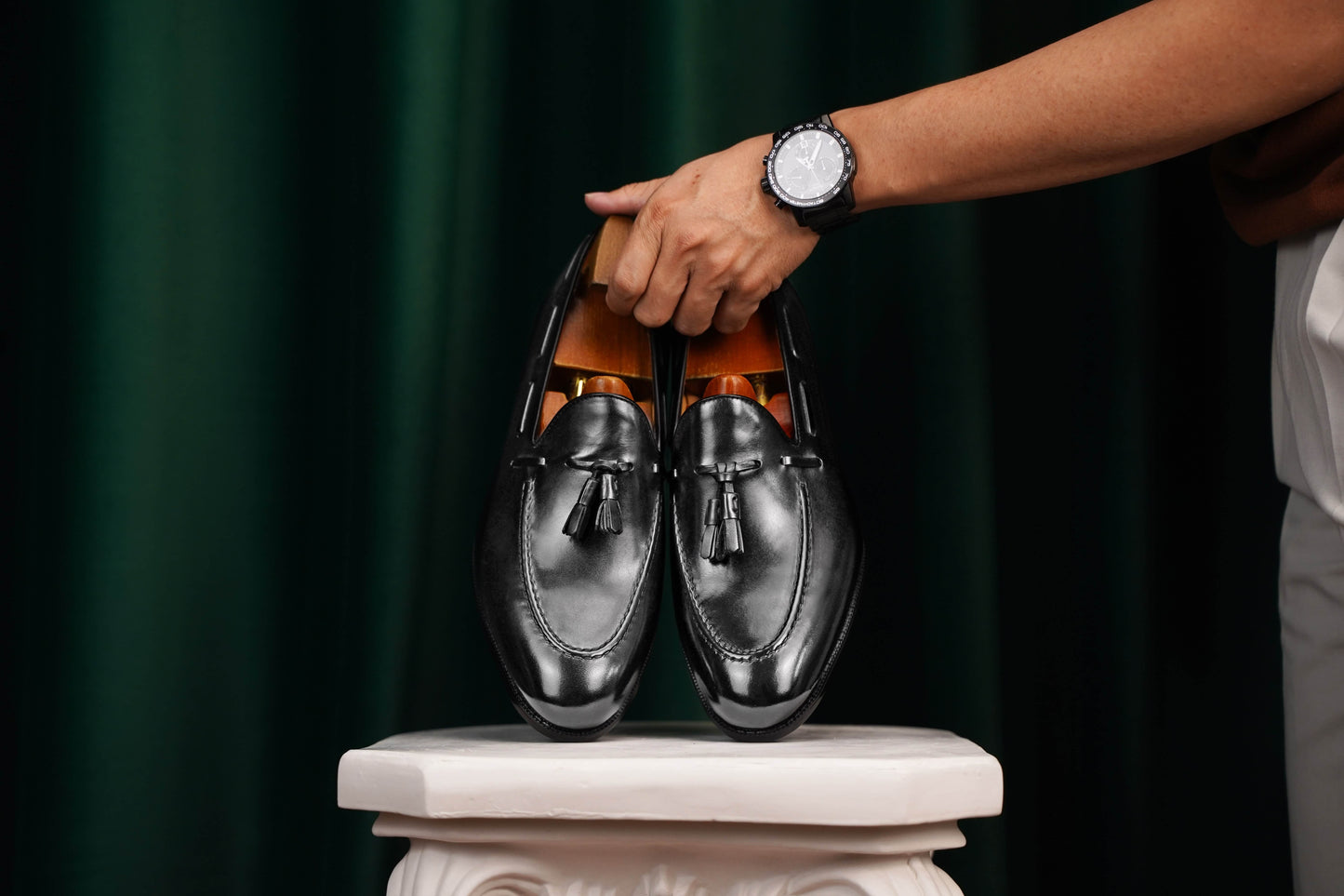 HolloShoe Handmade Black Tassel Leather Loafers – pair held by a man's hand with a watch, showcasing sleek black leather and classic design against a green curtain.