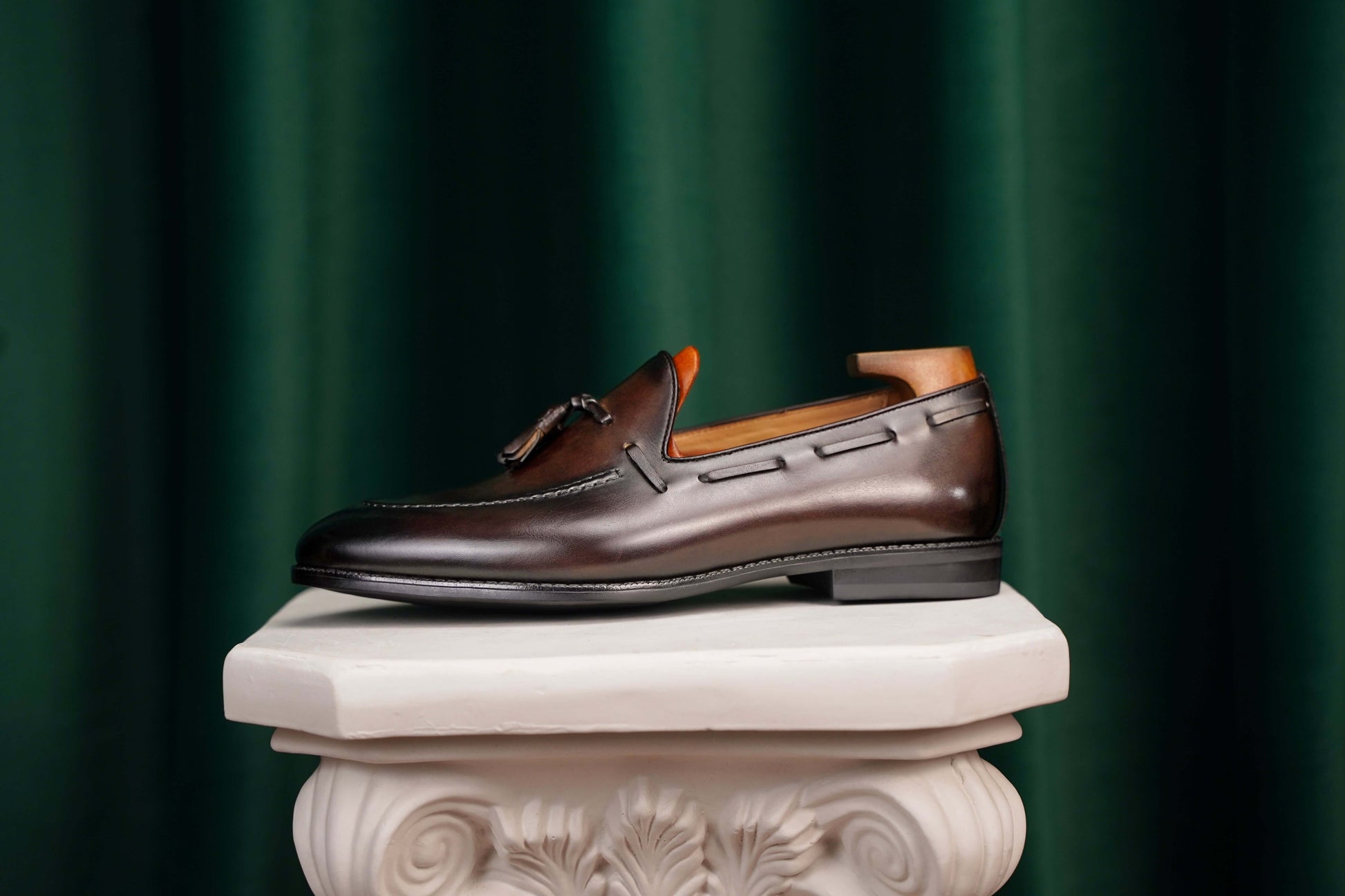 HolloShoe Handmade Chestnut Brown Patina Tassel Loafer – side view with shoe tree, resting on a classical pedestal, highlighting artisanal finish and refined style. 