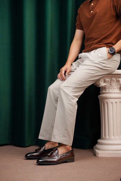 HolloShoe Handmade Chestnut Brown Patina Tassel Loafers – worn by a man in light trousers, sitting on a classical pedestal, highlighting artisanal finish and dressed-up ease.