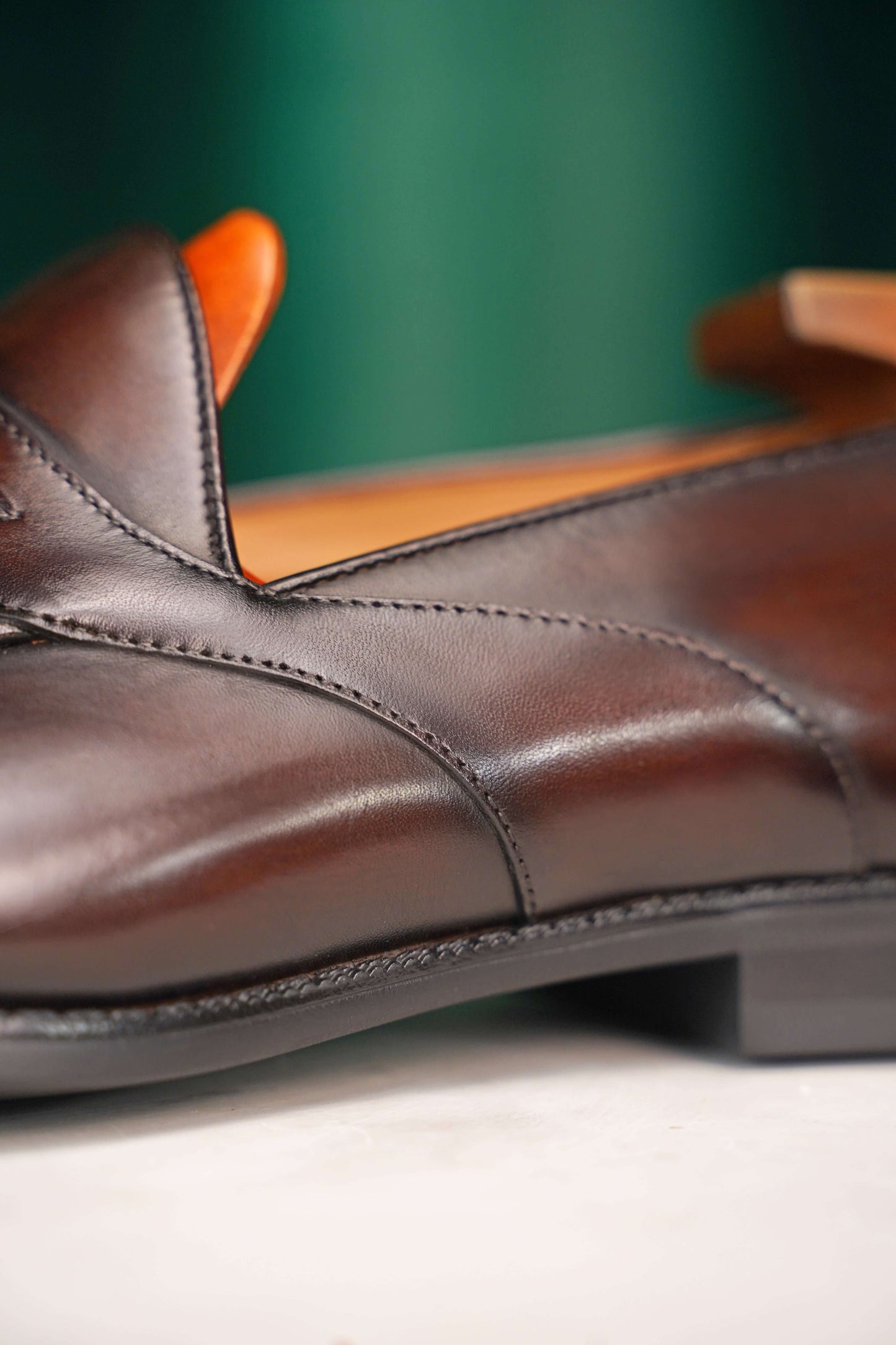 HolloShoe Handmade Dark Tan Leather Penny Loafers – side view with shoe tree, resting on a white classical pedestal against a green curtain, for elegant display.