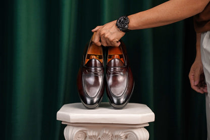 HolloShoe Handmade Dark Tan Leather Penny Loafers – pair held by a man's hand with a watch, highlighting sleek dark tan leather and classic design against a green curtain.