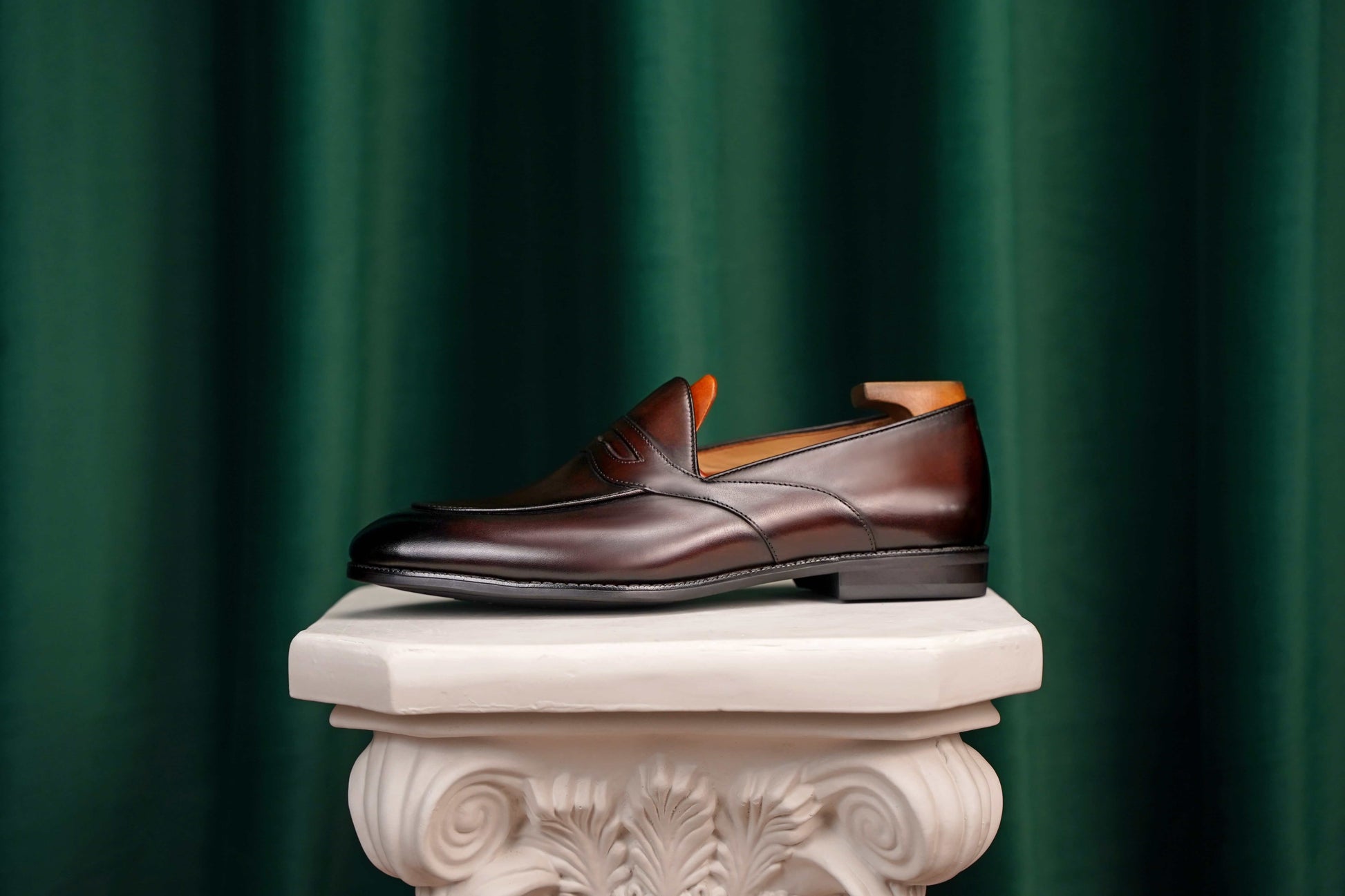 HolloShoe Handmade Dark Tan Leather Penny Loafers – side view with shoe tree, resting on a white classical pedestal against a green curtain, for elegant display.