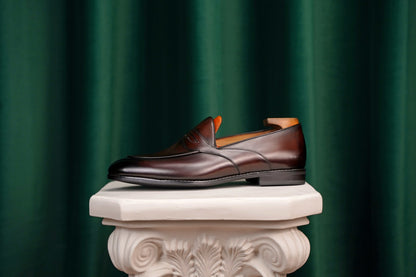 HolloShoe Handmade Dark Tan Leather Penny Loafers – side view with shoe tree, resting on a white classical pedestal against a green curtain, for elegant display.