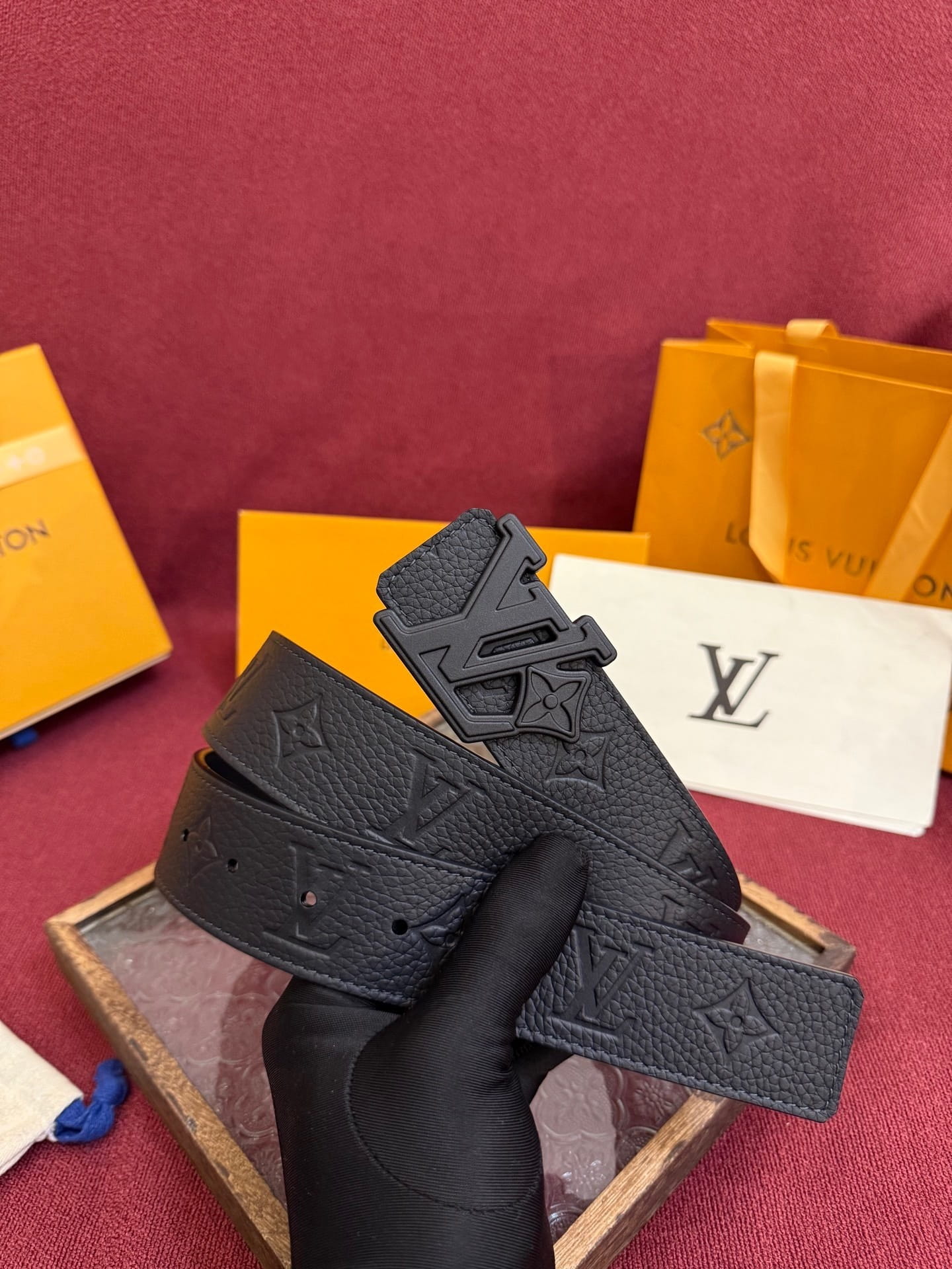 Black belt with a prominent buckle held by a gloved hand against a red background with Louis Vuitton packaging.