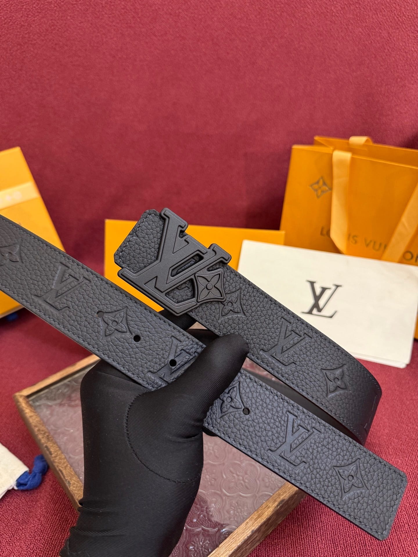 Louis Vuitton belt with monogram pattern held by a gloved hand against a red background.