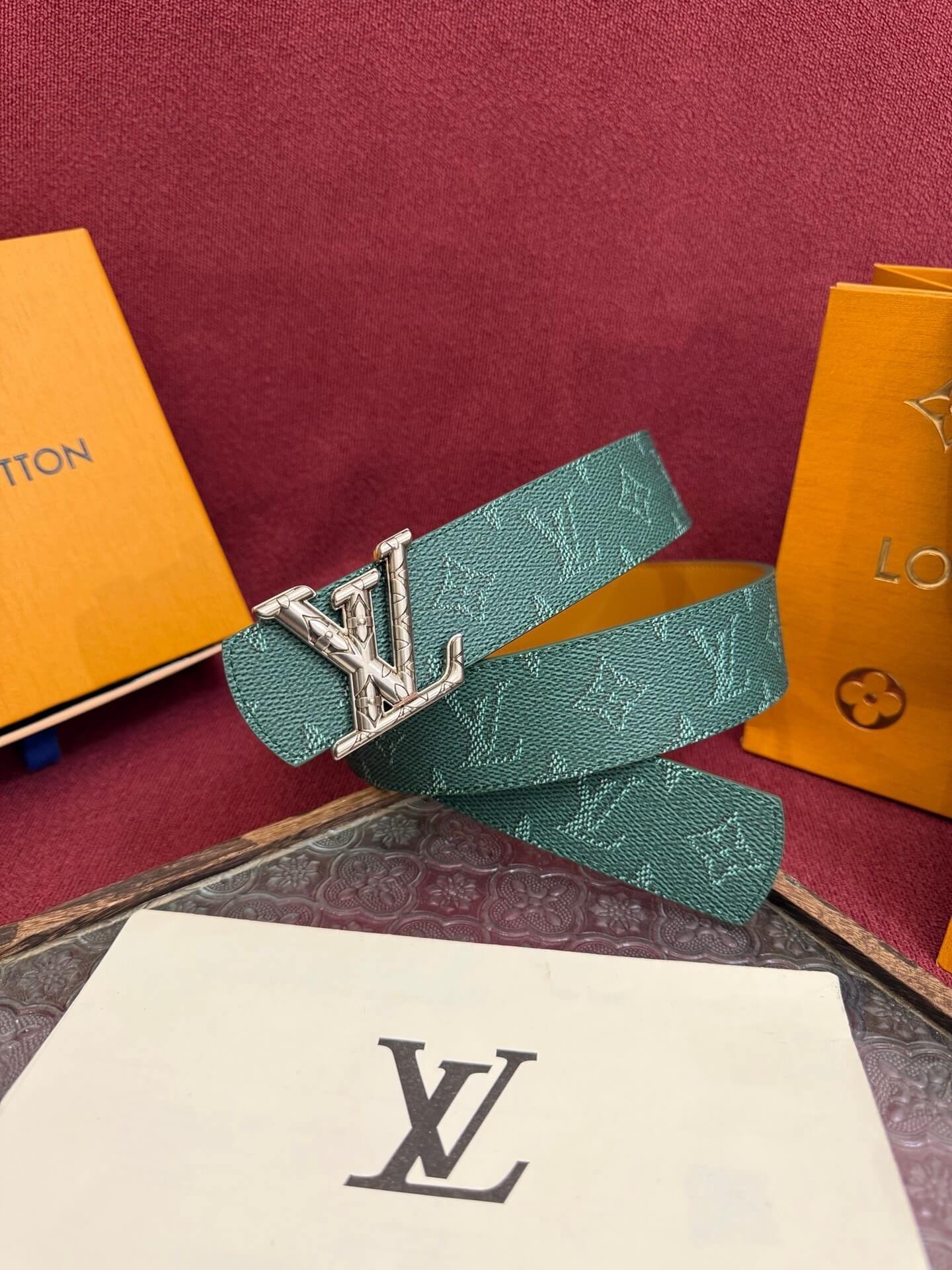 Green belt with silver buckle on a red surface with Louis Vuitton packaging.