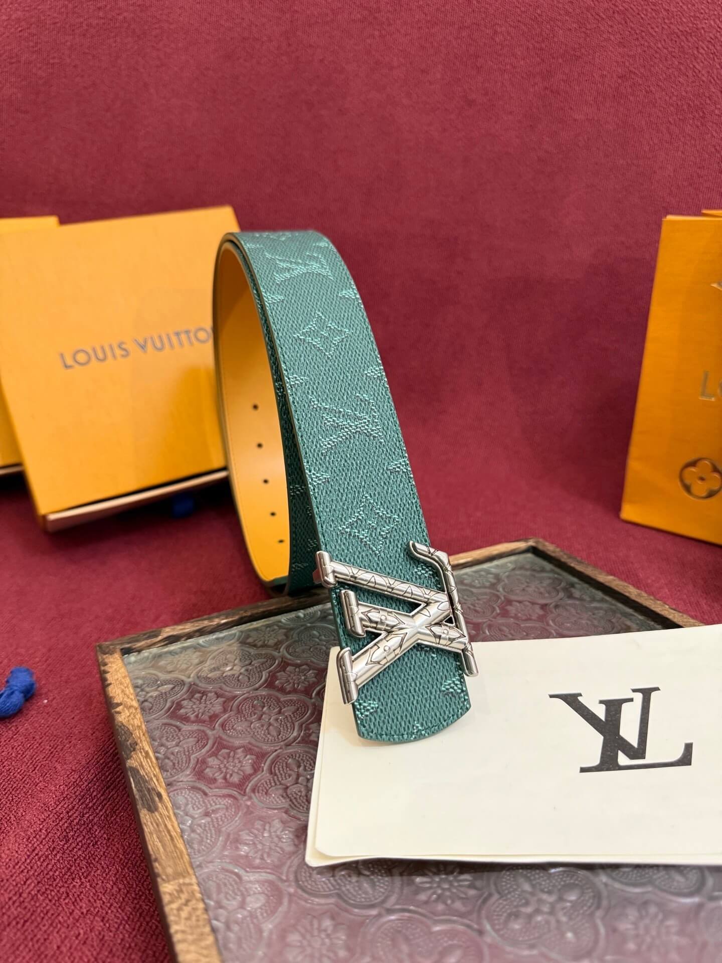 Teal Louis Vuitton belt with silver buckle on a red surface with brand packaging.