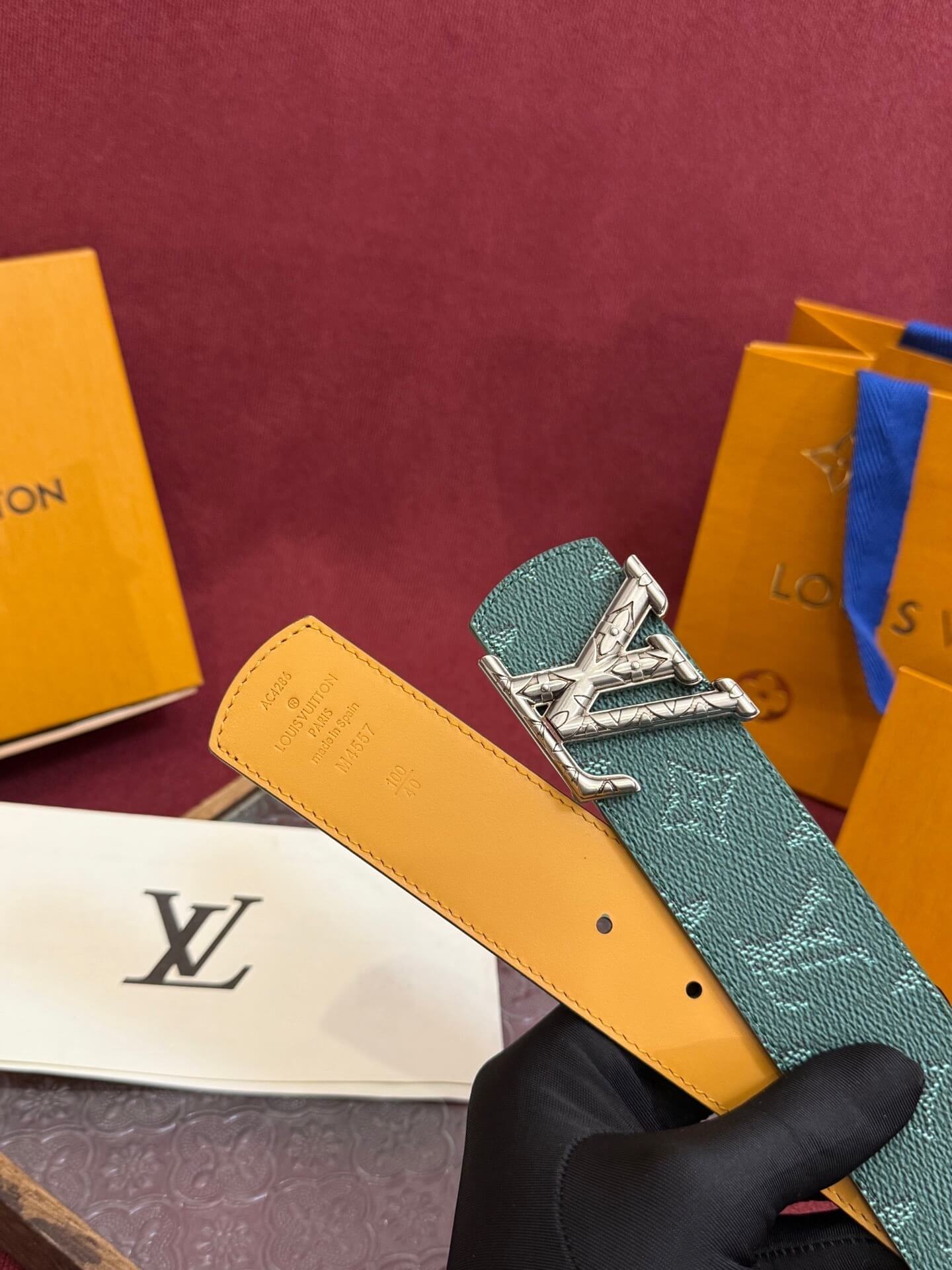 Teal Louis Vuitton belt with silver buckle held by a gloved hand against a maroon background with brand packaging.