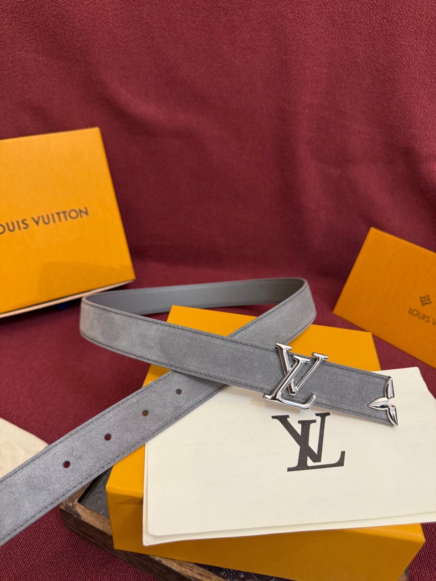 Gray belt with silver buckle on a red surface with Louis Vuitton packaging.