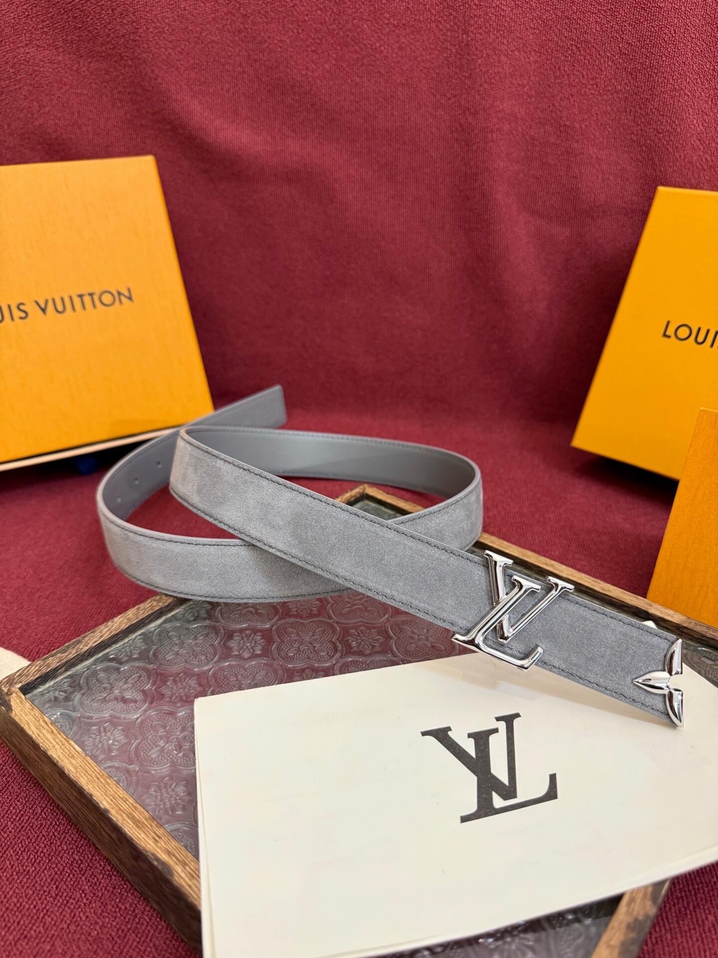 Gray belt with silver buckle on a wooden tray with Louis Vuitton packaging in the background.