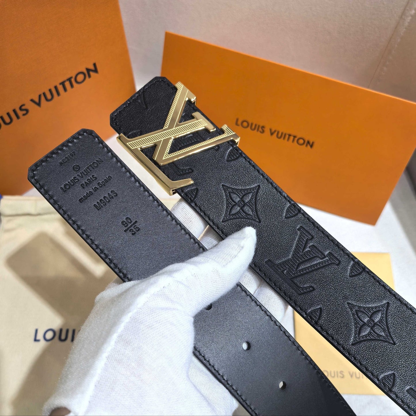 Black Louis Vuitton belt with gold buckle held by a gloved hand against orange Louis Vuitton boxes.