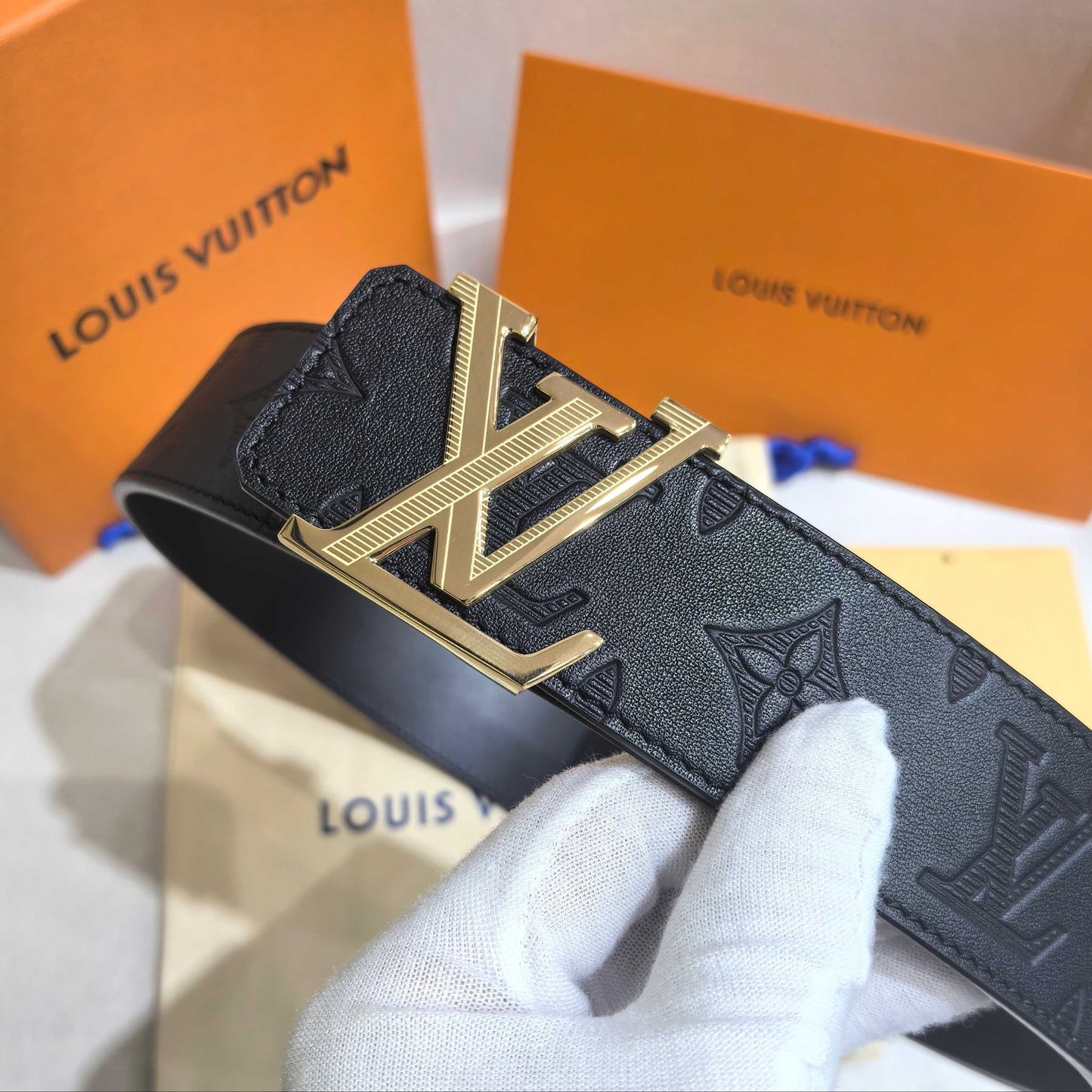 Black Louis Vuitton belt with gold buckle held by a gloved hand, with orange Louis Vuitton boxes in the background.