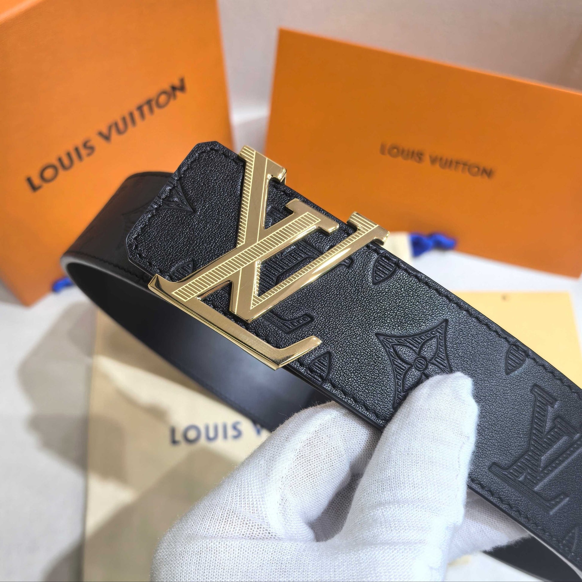 Black Louis Vuitton belt with gold buckle held by a gloved hand, with orange Louis Vuitton boxes in the background.
