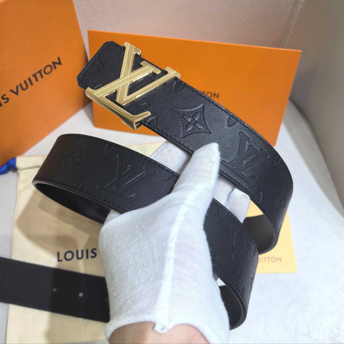 Black Louis Vuitton belt with gold buckle held by a gloved hand, with brand packaging in the background.