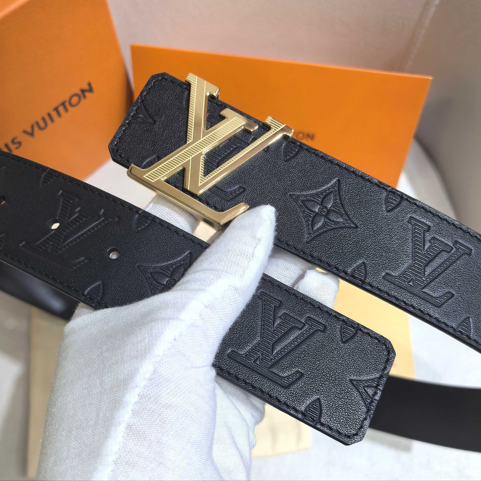 Louis Vuitton belt with gold buckle held by a gloved hand, with an orange Louis Vuitton box in the background.