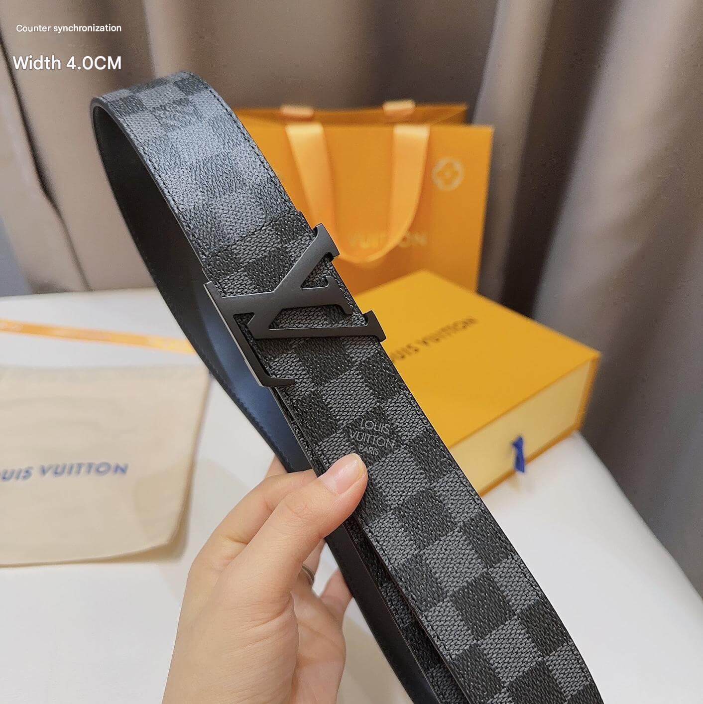 Louis Vuitton belt with a hand holding it, with a blurred background of a Louis Vuitton box and paper.
