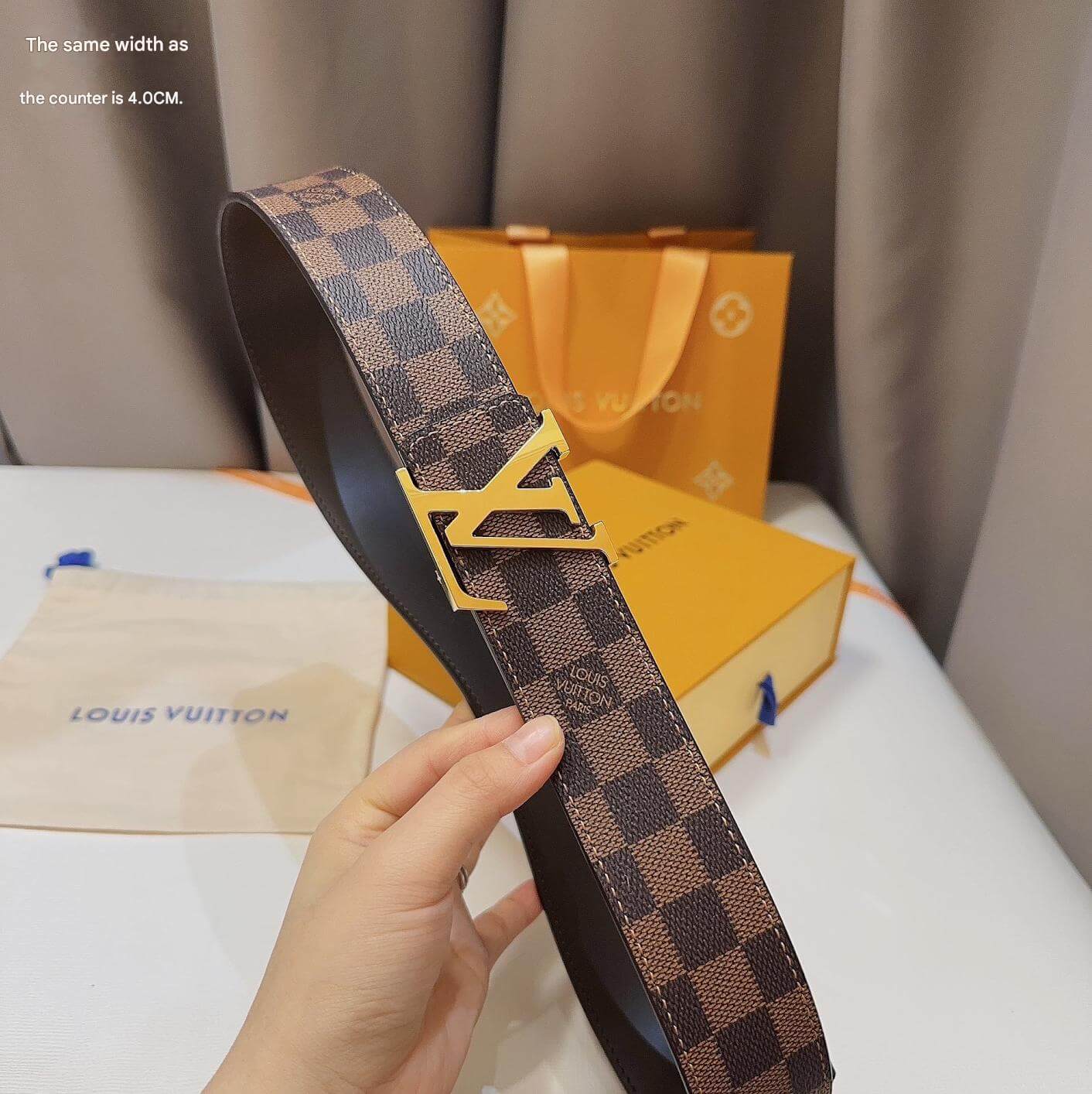 Louis Vuitton belt with checkered pattern and gold buckle held by a hand, with a Louis Vuitton box and pouch in the background.