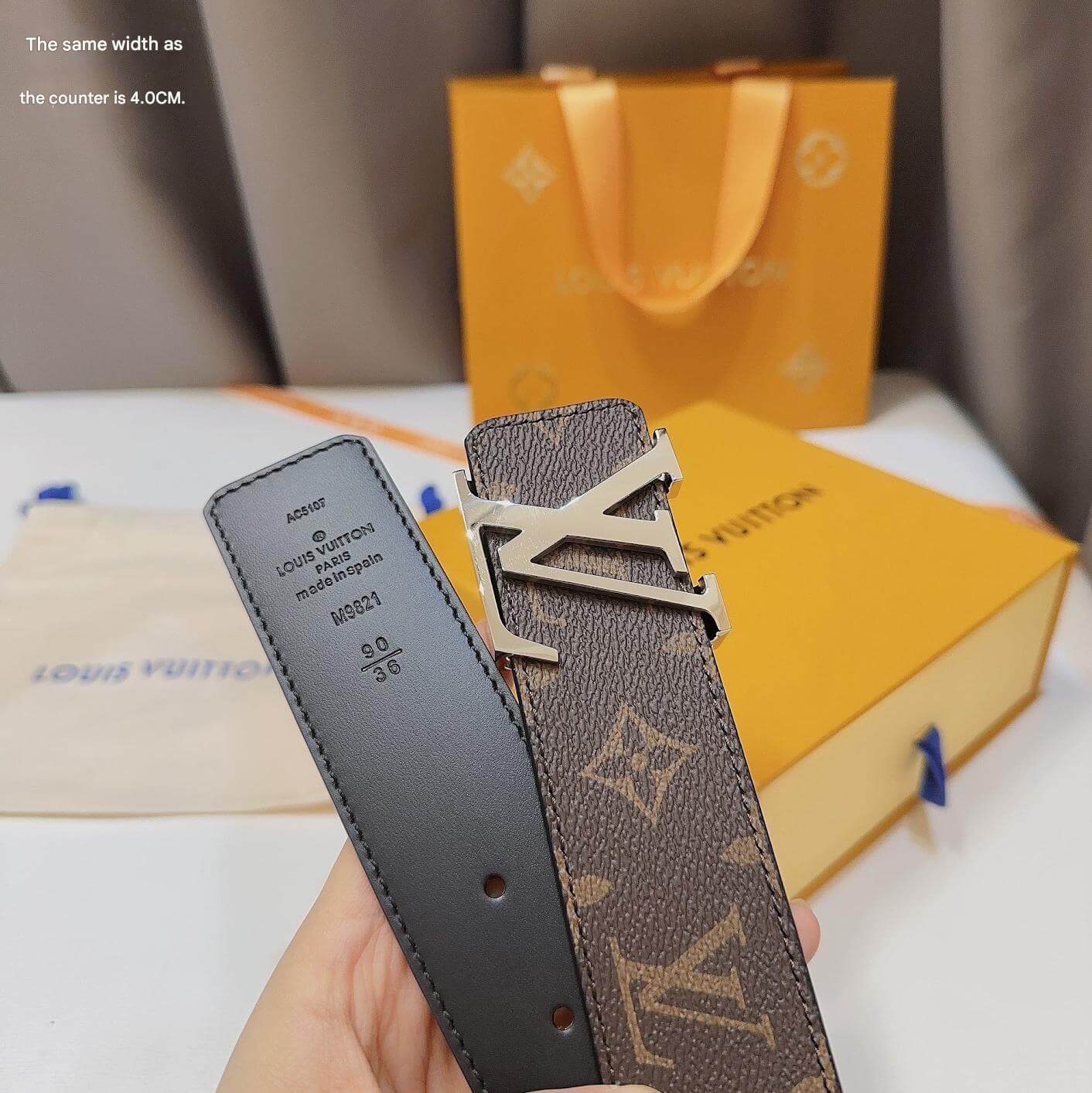 Louis Vuitton belt with monogram pattern held by a hand, with a yellow Louis Vuitton box in the background.