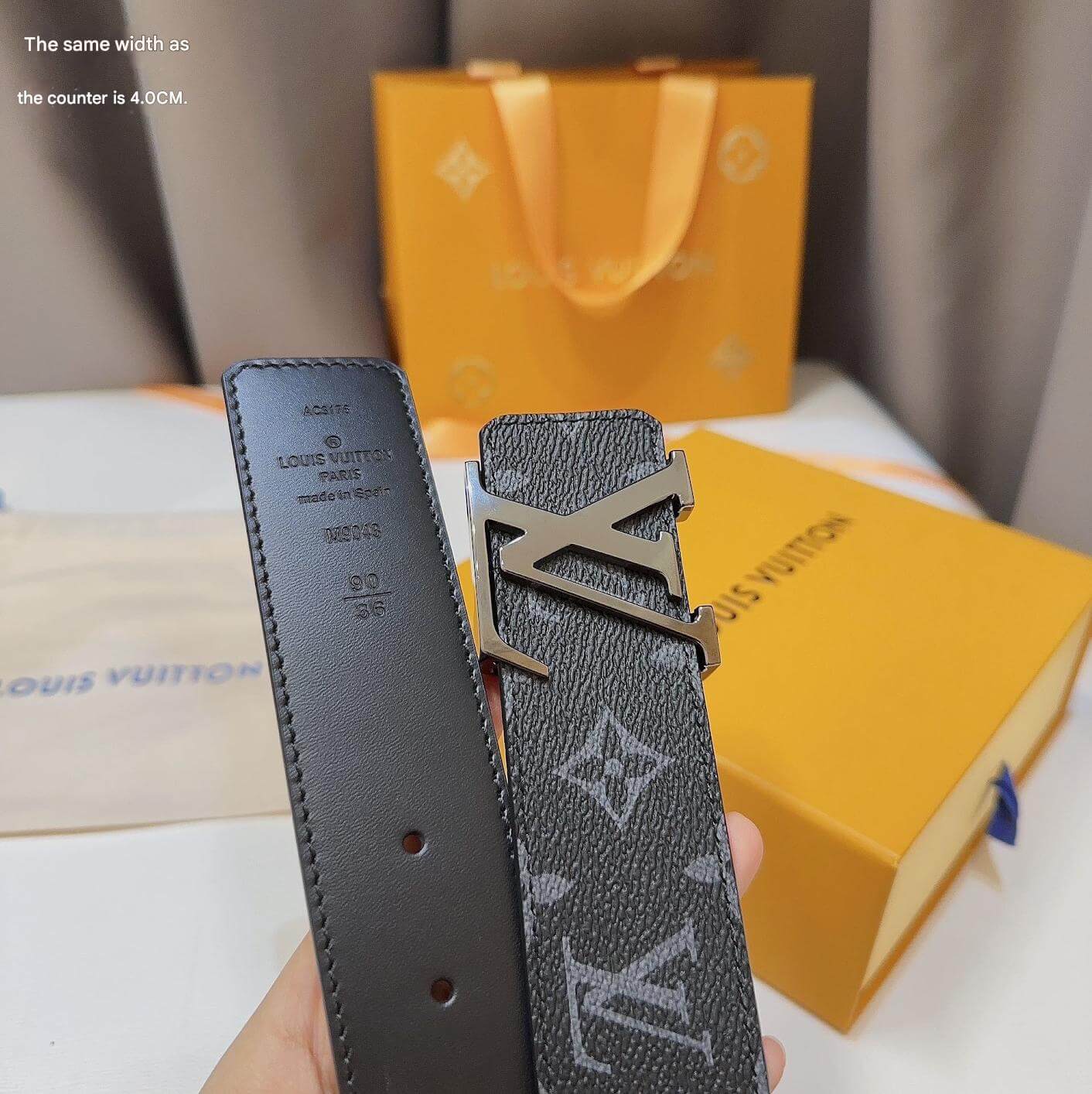 Louis Vuitton belt with monogram pattern held in front of a yellow box.