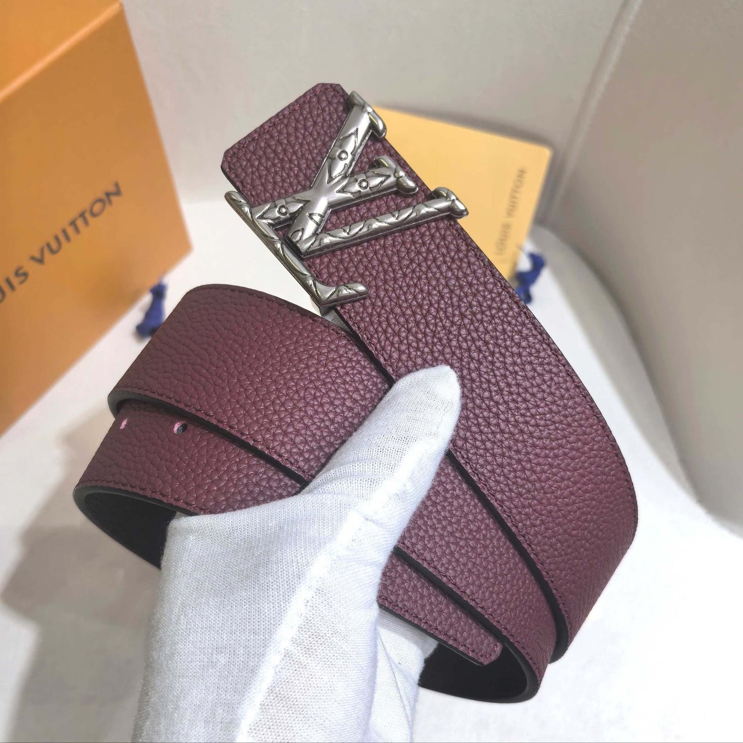 Maroon belt with silver buckle held by a white gloved hand, with a Louis Vuitton box in the background.