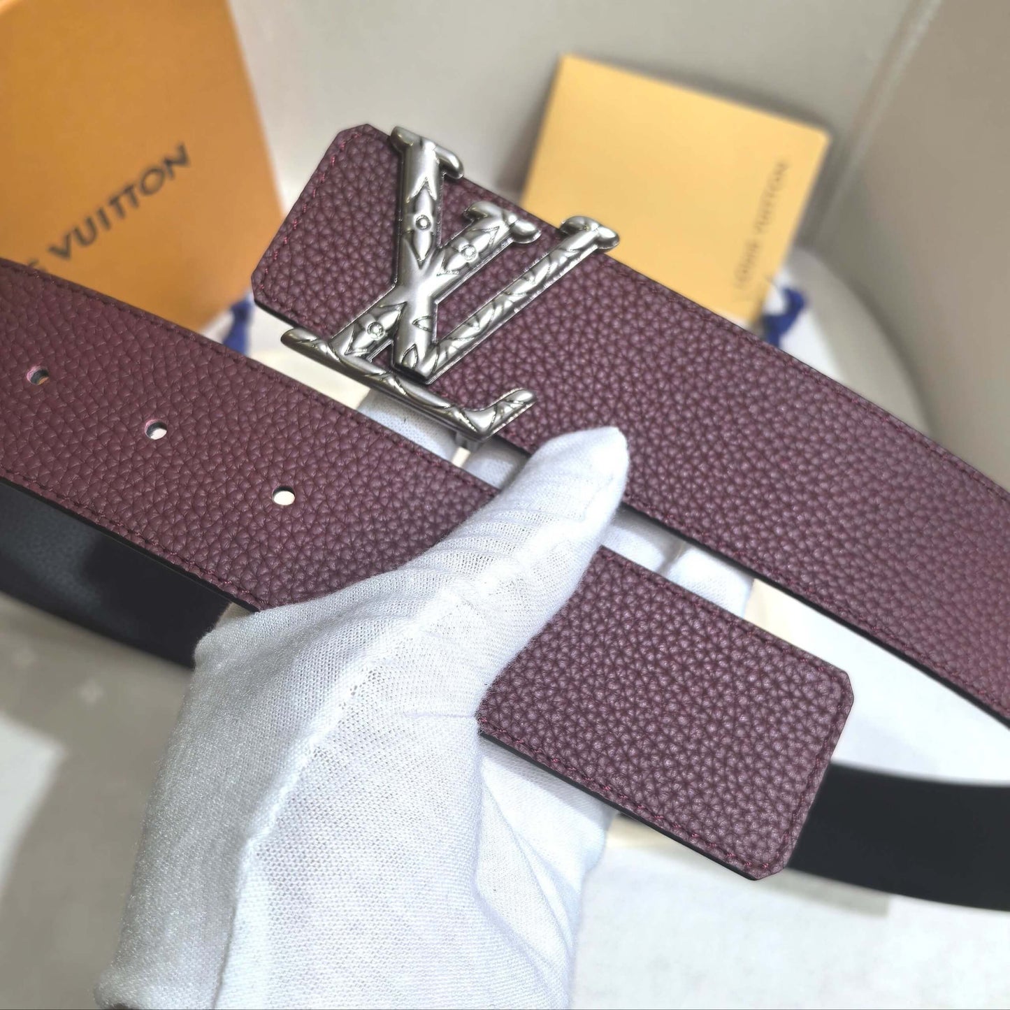 Maroon belt with silver buckle held by a gloved hand, with 'Louis Vuitton' branding in the background.
