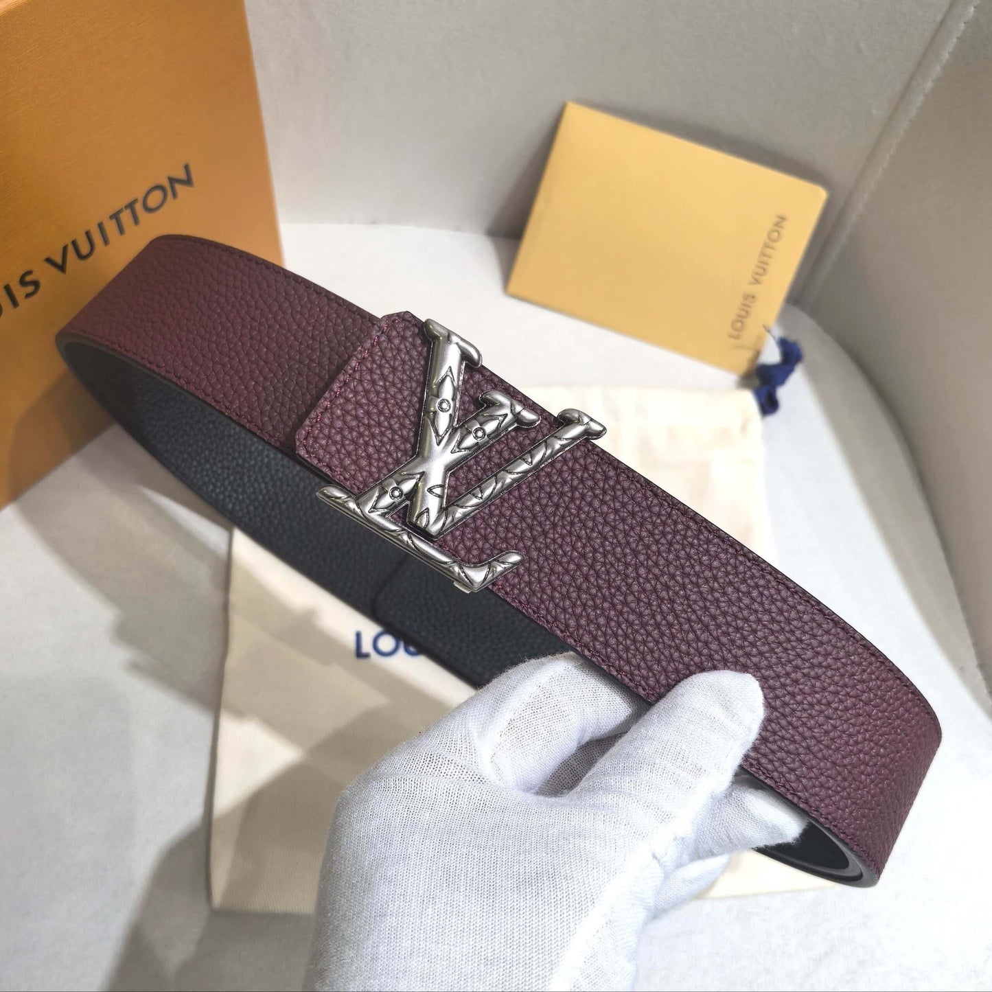 Maroon belt with silver buckle held by a gloved hand, with Louis Vuitton boxes in the background.