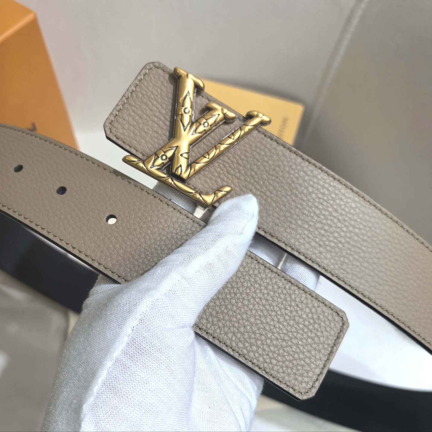 Beige belt with gold buckle held by a gloved hand against a neutral background