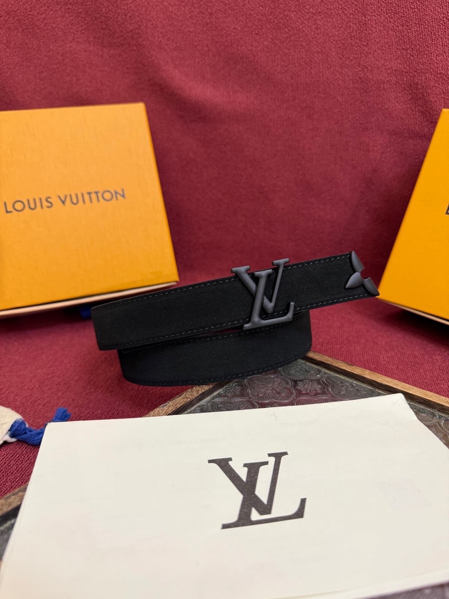 Black Louis Vuitton belt with logo on a red surface with brand boxes in the background.