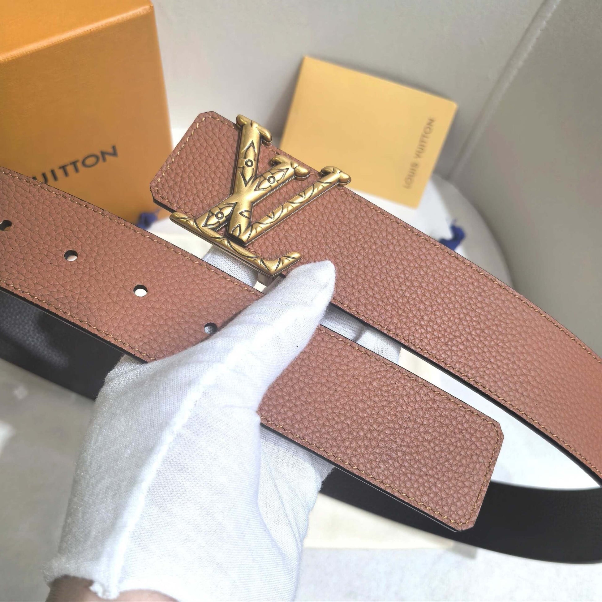 Brown belt with gold buckle on a white surface, with a Louis Vuitton box in the background.