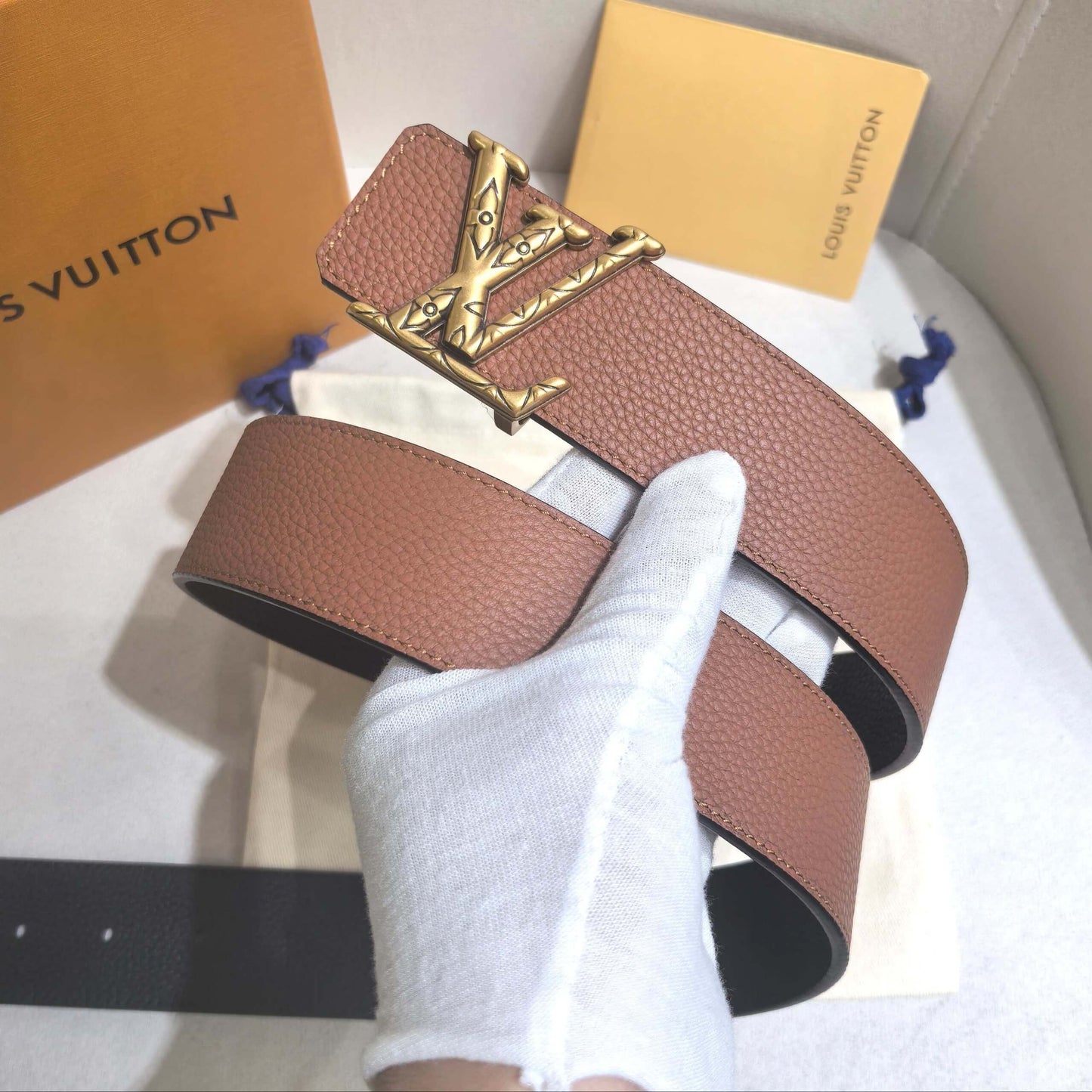 Brown leather belt with gold buckle held by a gloved hand on a white surface with Louis Vuitton branding.