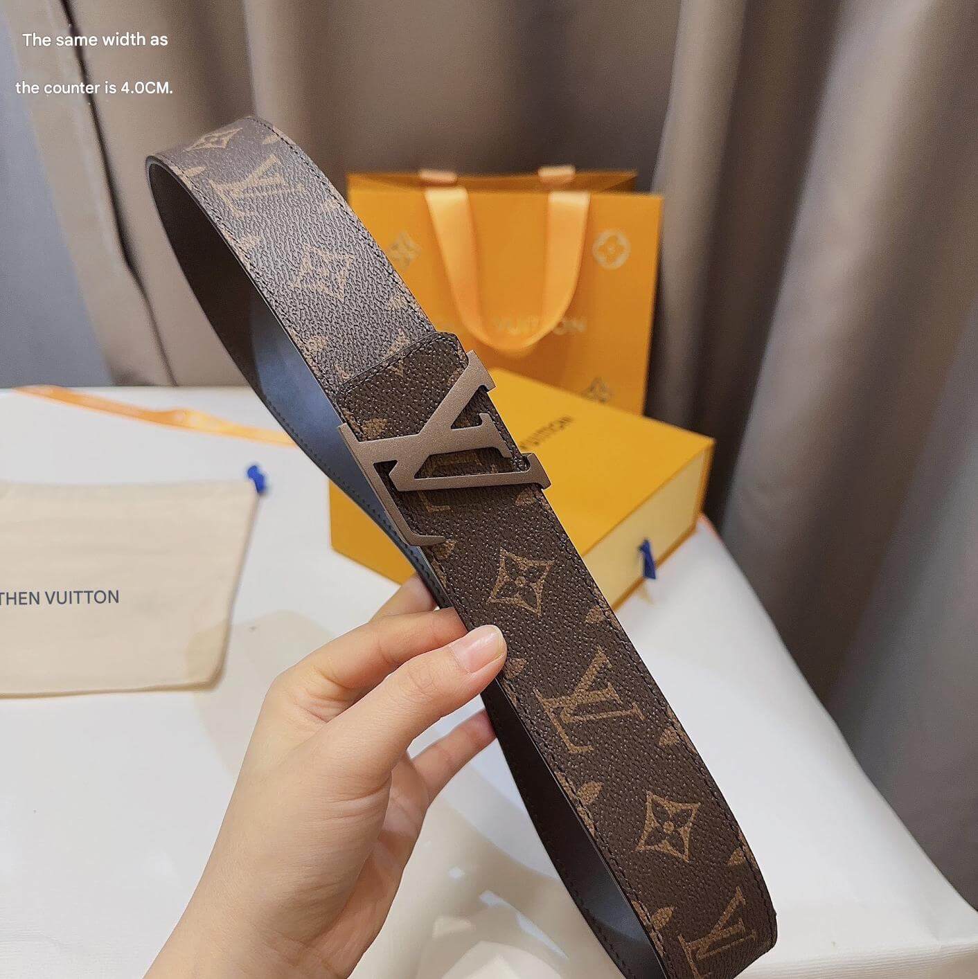 Brown belt with monogram pattern held by a hand, with a blurred background of a box and packaging.