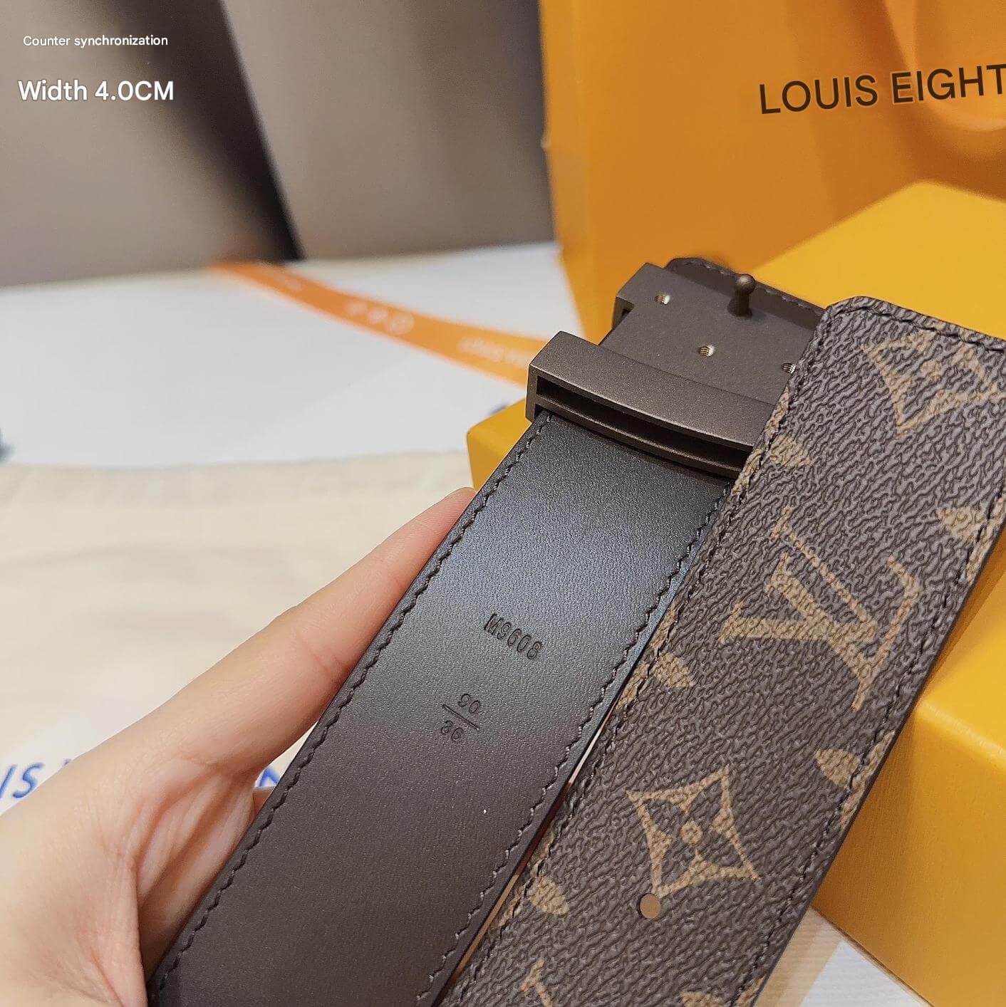 Louis Vuitton belt with monogram pattern held by a hand, with a yellow box in the background.