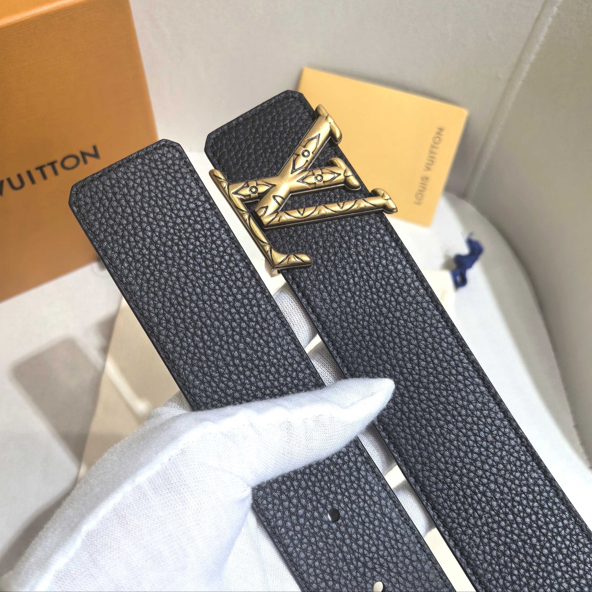 Black belt with gold buckle held by a gloved hand, with Louis Vuitton box and paper in the background.