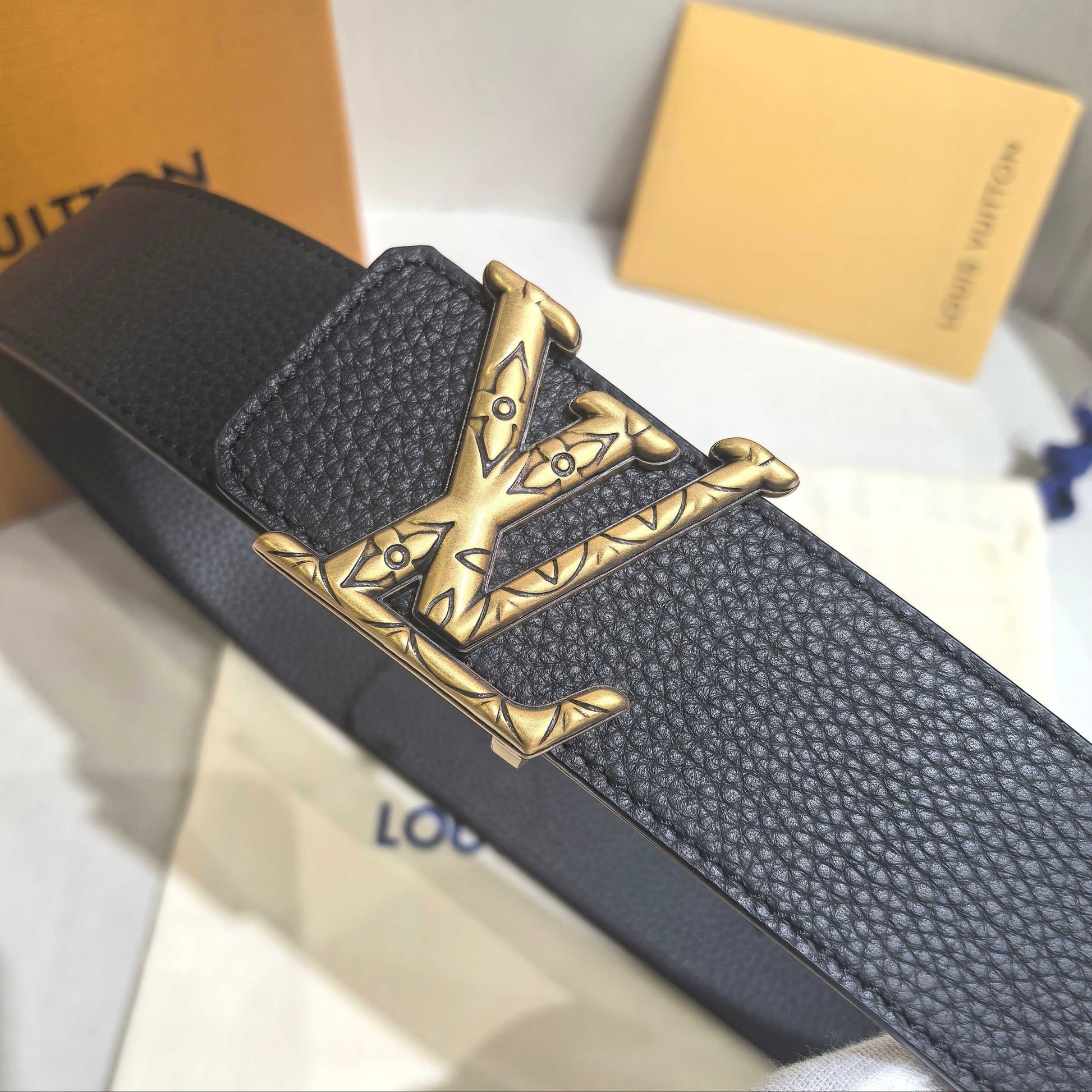 Black leather belt with gold buckle on a white surface, with Louis Vuitton packaging in the background.