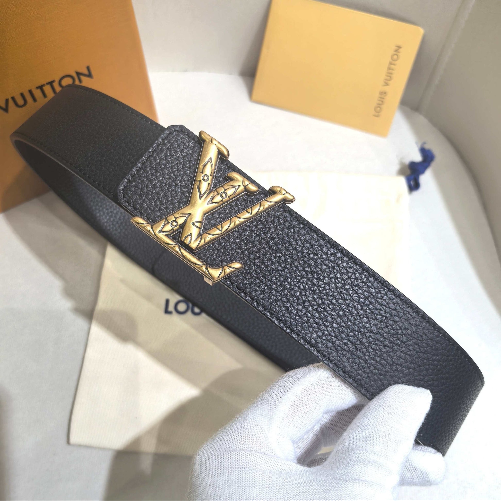 Black leather belt with gold buckle held by a gloved hand, with Louis Vuitton packaging in the background.