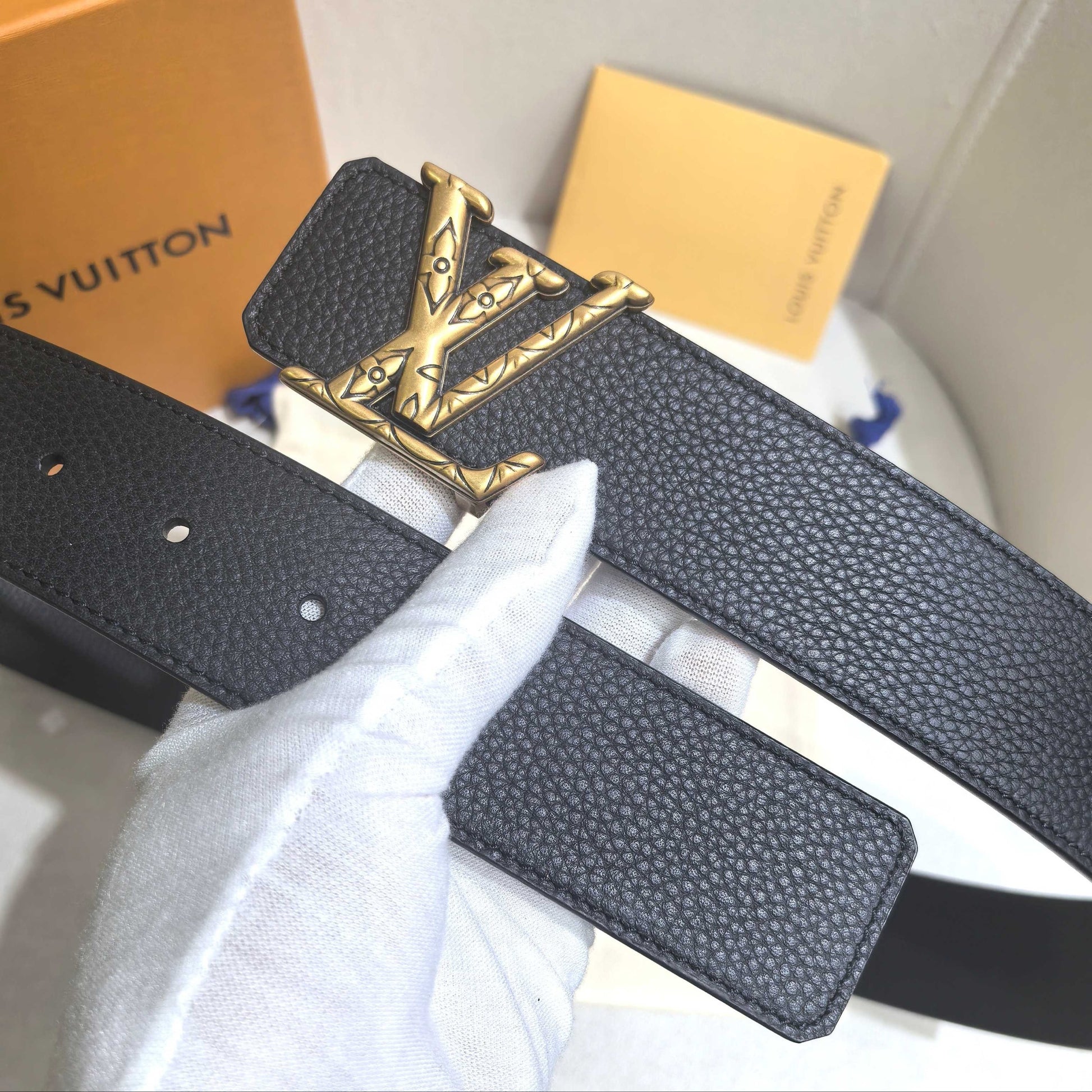 Black leather belt with gold buckle held by a gloved hand, with Louis Vuitton packaging in the background.