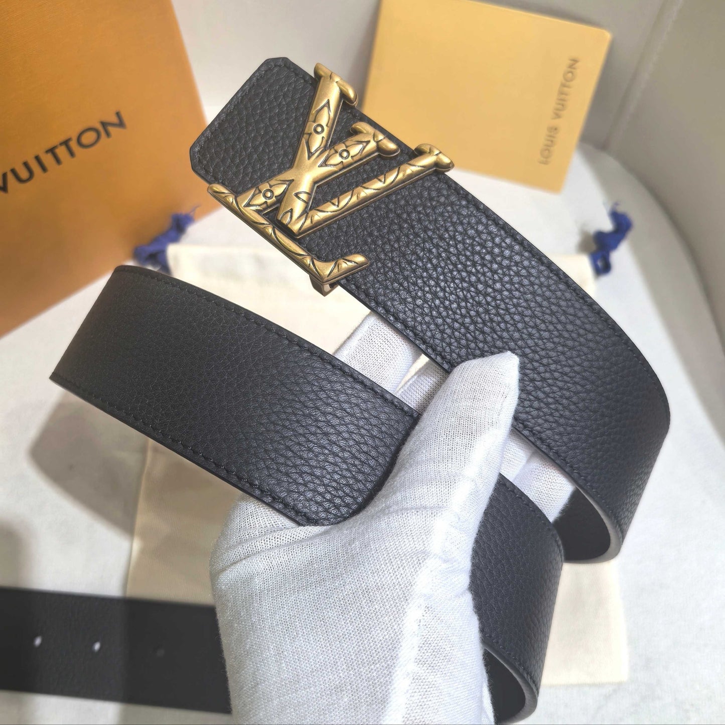 Black leather belt with gold buckle held by a gloved hand, with Louis Vuitton packaging in the background.