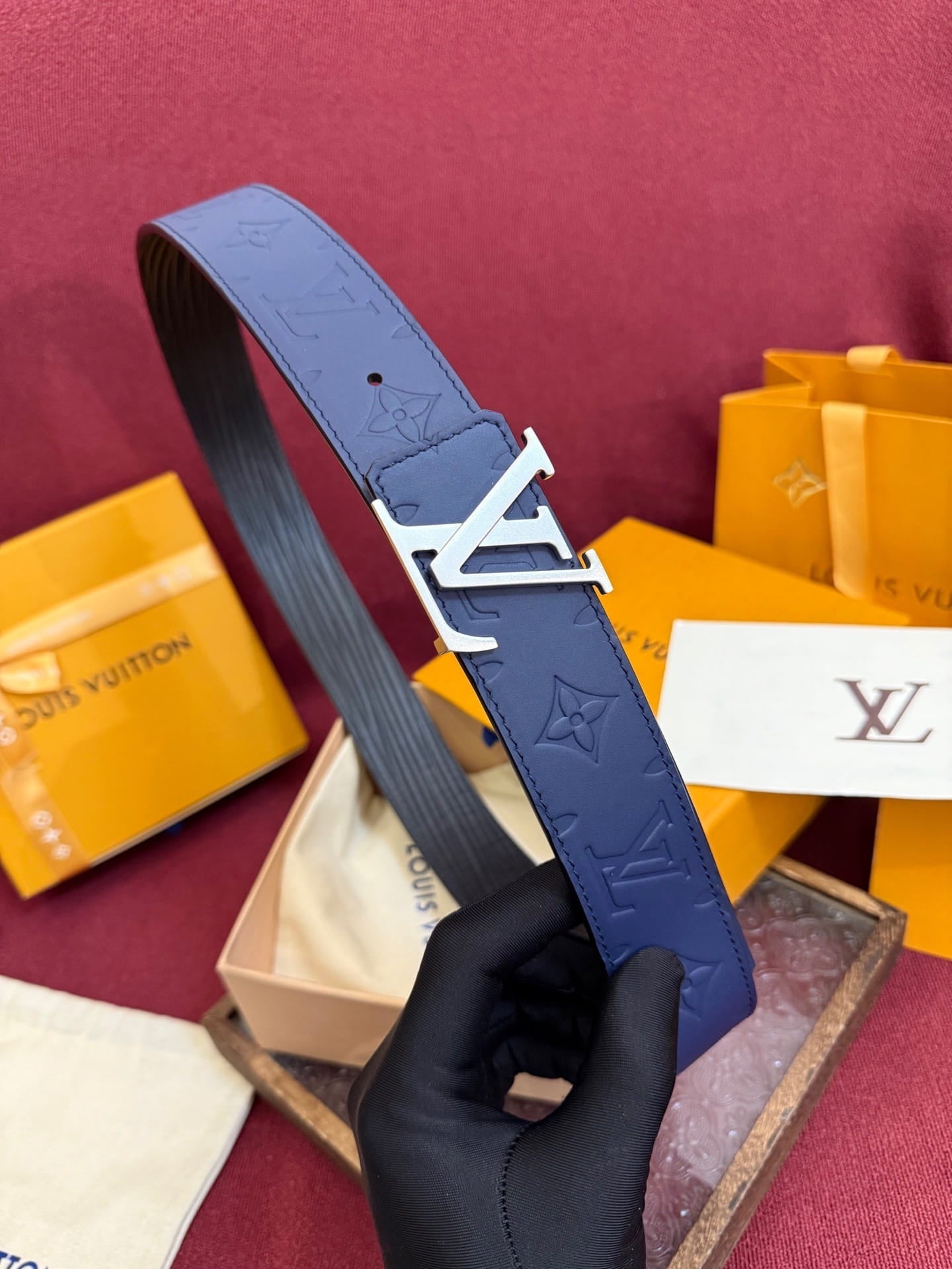 Blue belt with silver buckle held by a gloved hand against a maroon background with Louis Vuitton packaging.