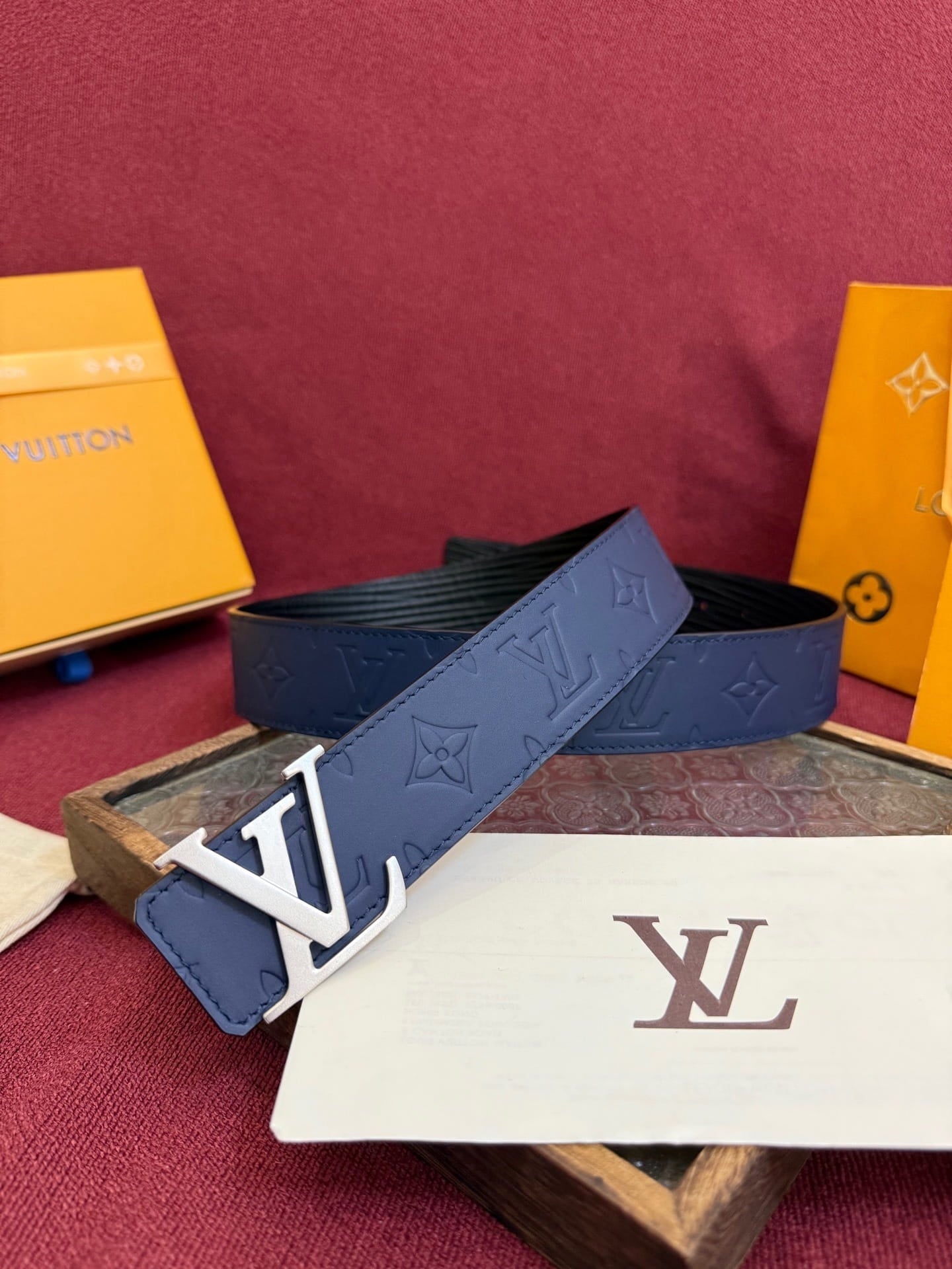 Blue belt with monogram pattern and white logo on a red surface with Louis Vuitton boxes.