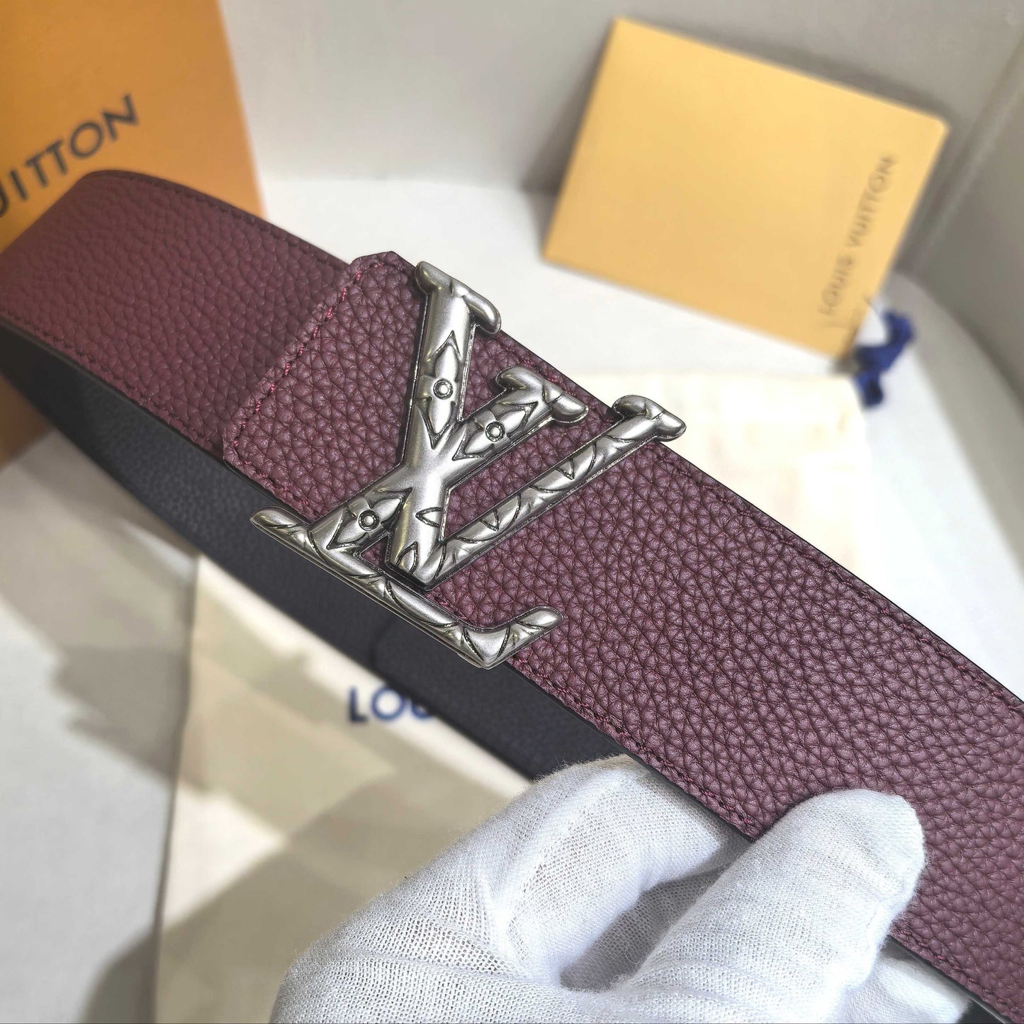 Maroon belt with silver buckle held by a gloved hand, with Louis Vuitton packaging in the background.
