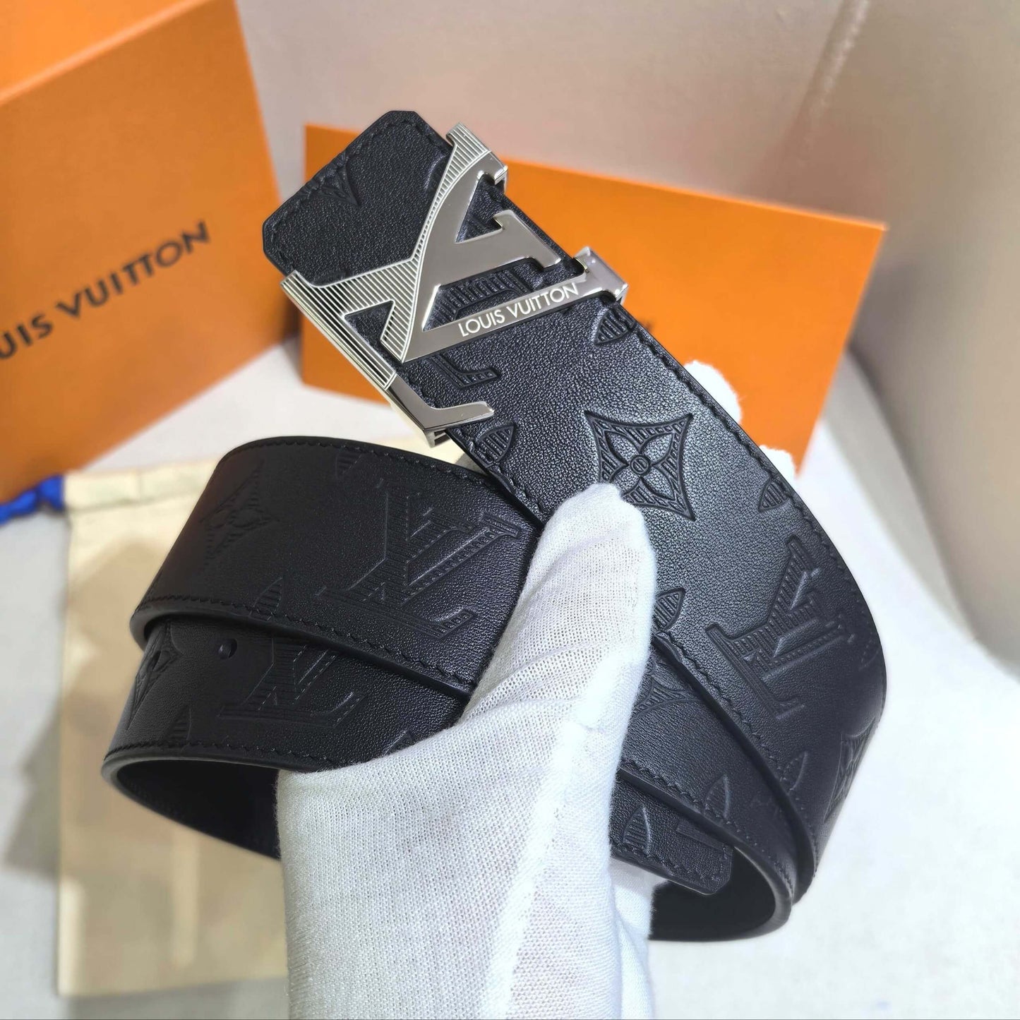 Black Louis Vuitton belt with silver buckle held by a gloved hand, with an orange Louis Vuitton box in the background.