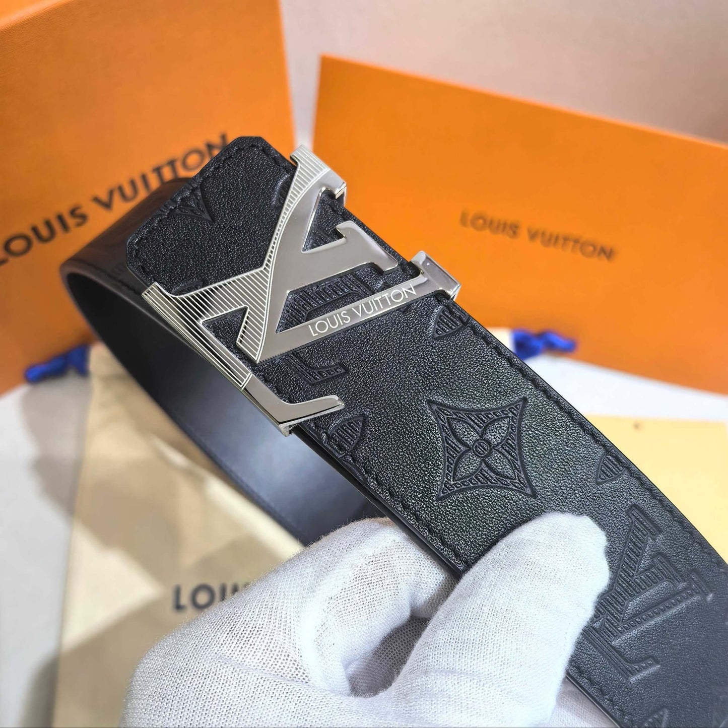 Louis Vuitton belt with silver buckle held by a gloved hand against an orange Louis Vuitton box.