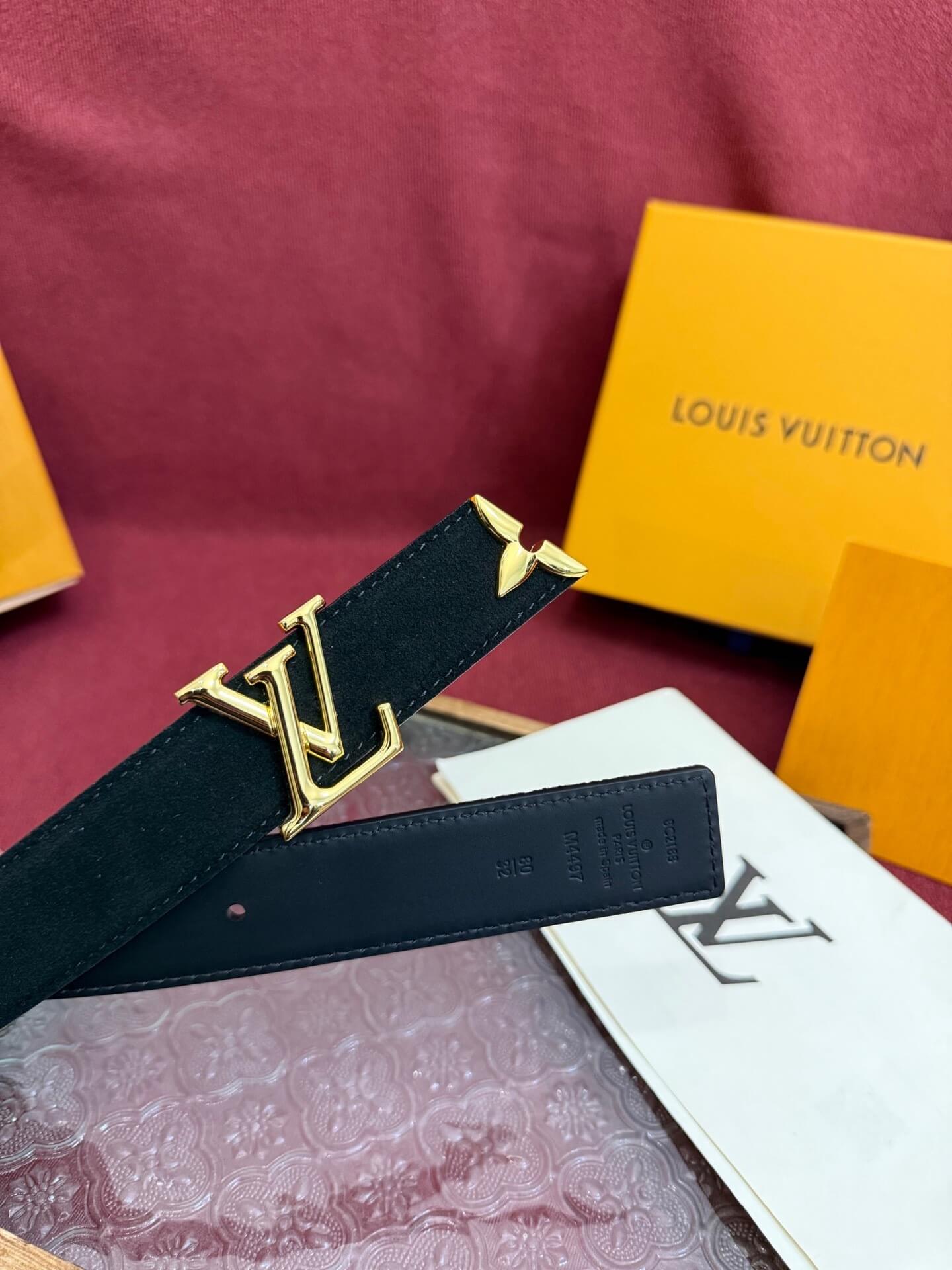 Black belt with gold buckle on a maroon surface with Louis Vuitton packaging.