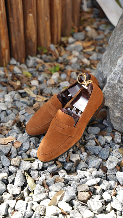 Luxurious Sand Suede Penny Loafers – pair resting on gravel and rocks, emphasizing sand-colored suede texture and iconic penny loafer design.