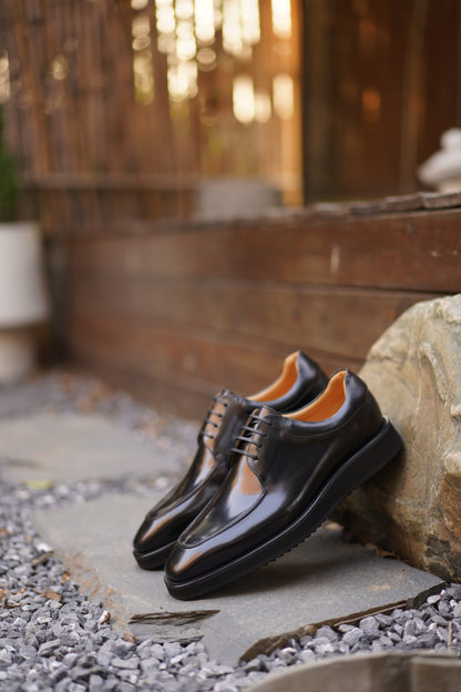Pair of Black Apron-Toe Derby shoes with high-shine black leather, elevated lug soles, and shoe trees, angled view.