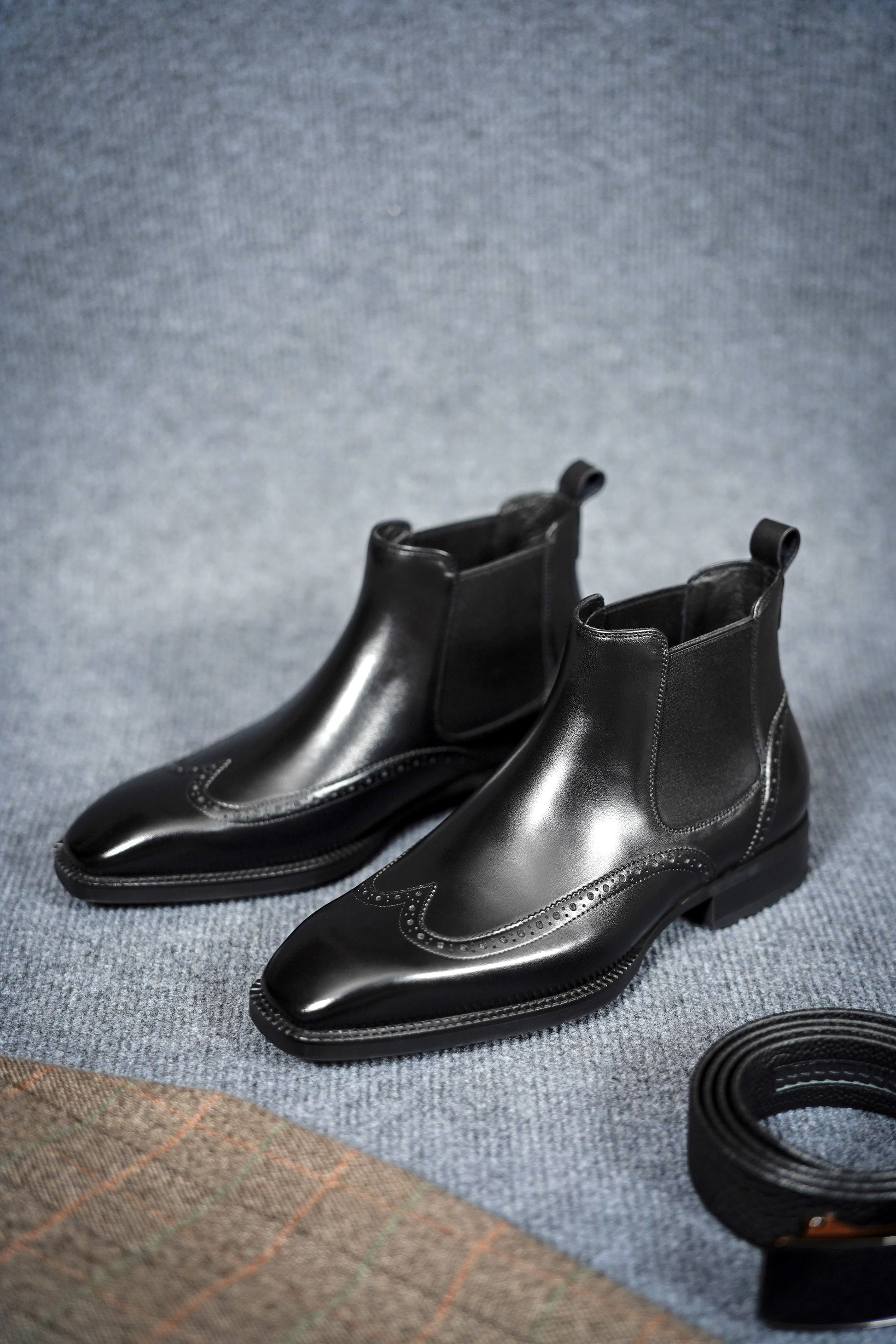 Modern Black Brogue Chelsea Boots for Sophisticated Style – a pair of black leather boots on a gray textured background next to a belt, showcasing their elegant design.