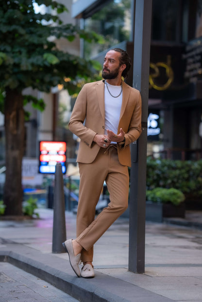 A stylish man wearing a Camel Mono Collar Slim-Fit Suit stands confidently in front of a modern storefront. The suit is paired with a simple white T-shirt, creating a relaxed yet refined look. He accessorizes with a chain necklace, a ring, and a watch, while white tassel loafers complete the ensemble. The man has a neatly groomed beard and his hair pulled back, exuding a contemporary and sophisticated vibe, perfectly balancing casual and formal elements in his outfit.