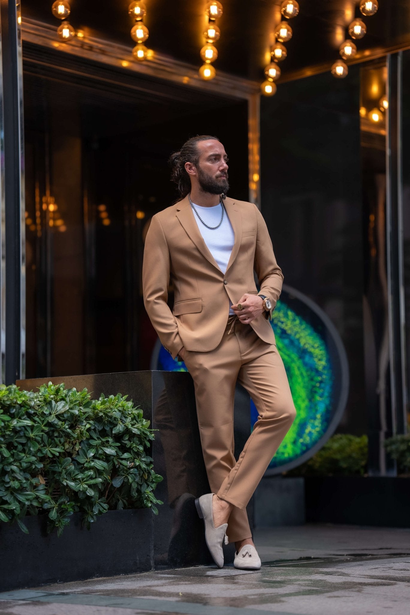 A stylish man wearing a Camel Mono Collar Slim-Fit Suit stands confidently in front of a modern storefront. The suit is paired with a simple white T-shirt, creating a relaxed yet refined look. He accessorizes with a chain necklace, a ring, and a watch, while white tassel loafers complete the ensemble. The man has a neatly groomed beard and his hair pulled back, exuding a contemporary and sophisticated vibe, perfectly balancing casual and formal elements in his outfit.